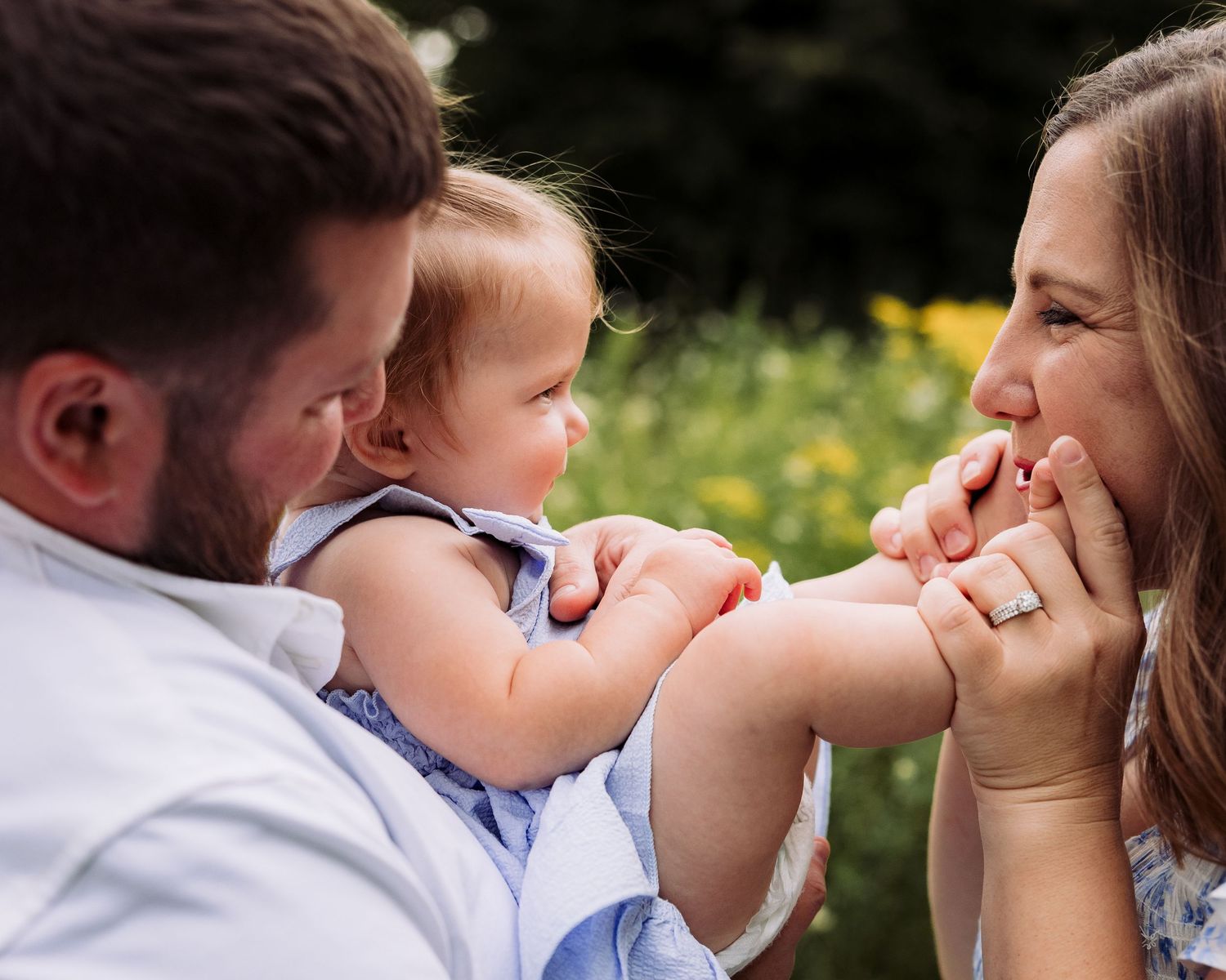 A couple smiles at their baby outdoors, surrounded by greenery and yellow flowers, sharing a joyful family moment.