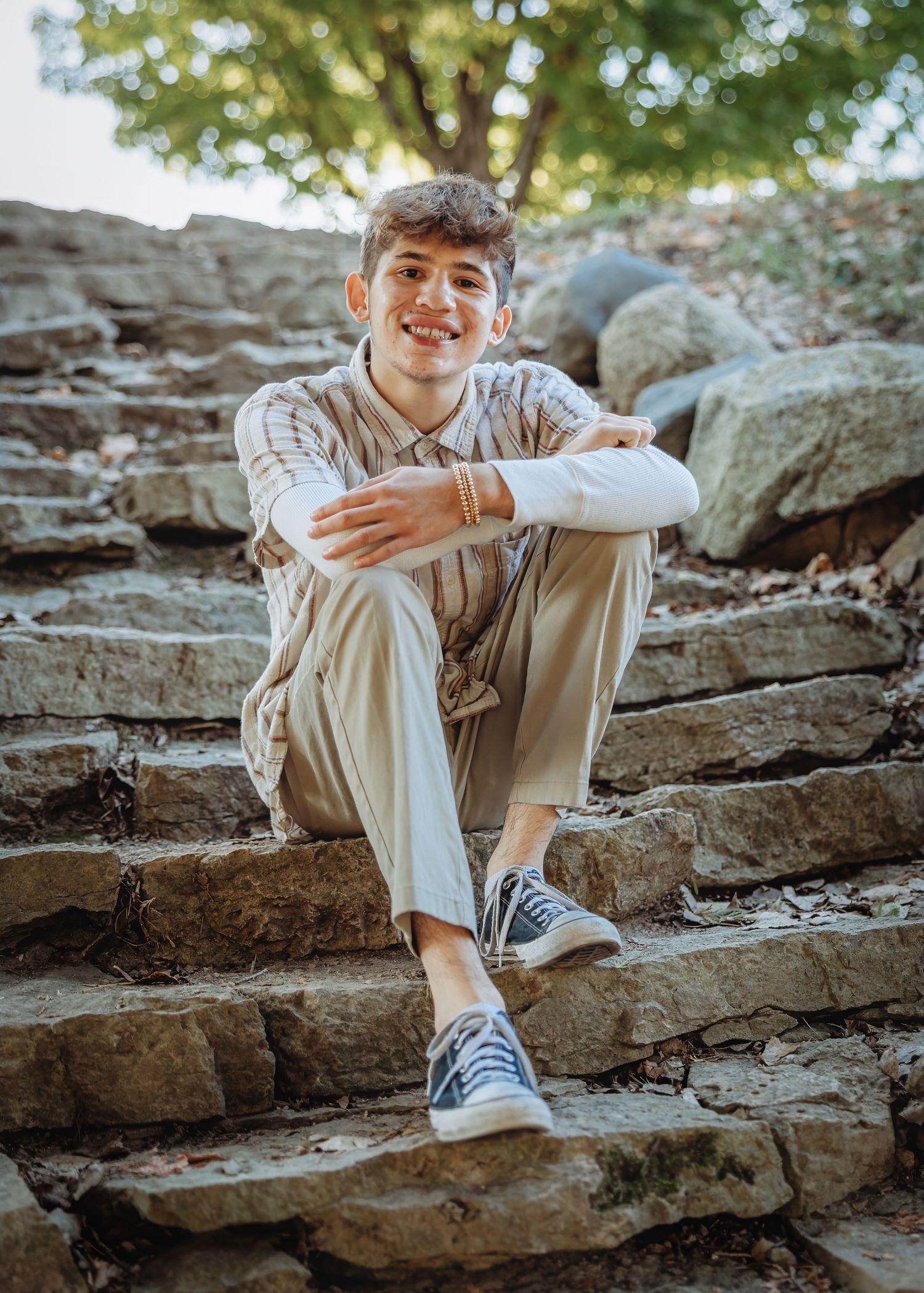 Smiling teenage boy sitting on stone steps outdoors, wearing striped shirt, beige pants, and blue sneakers.