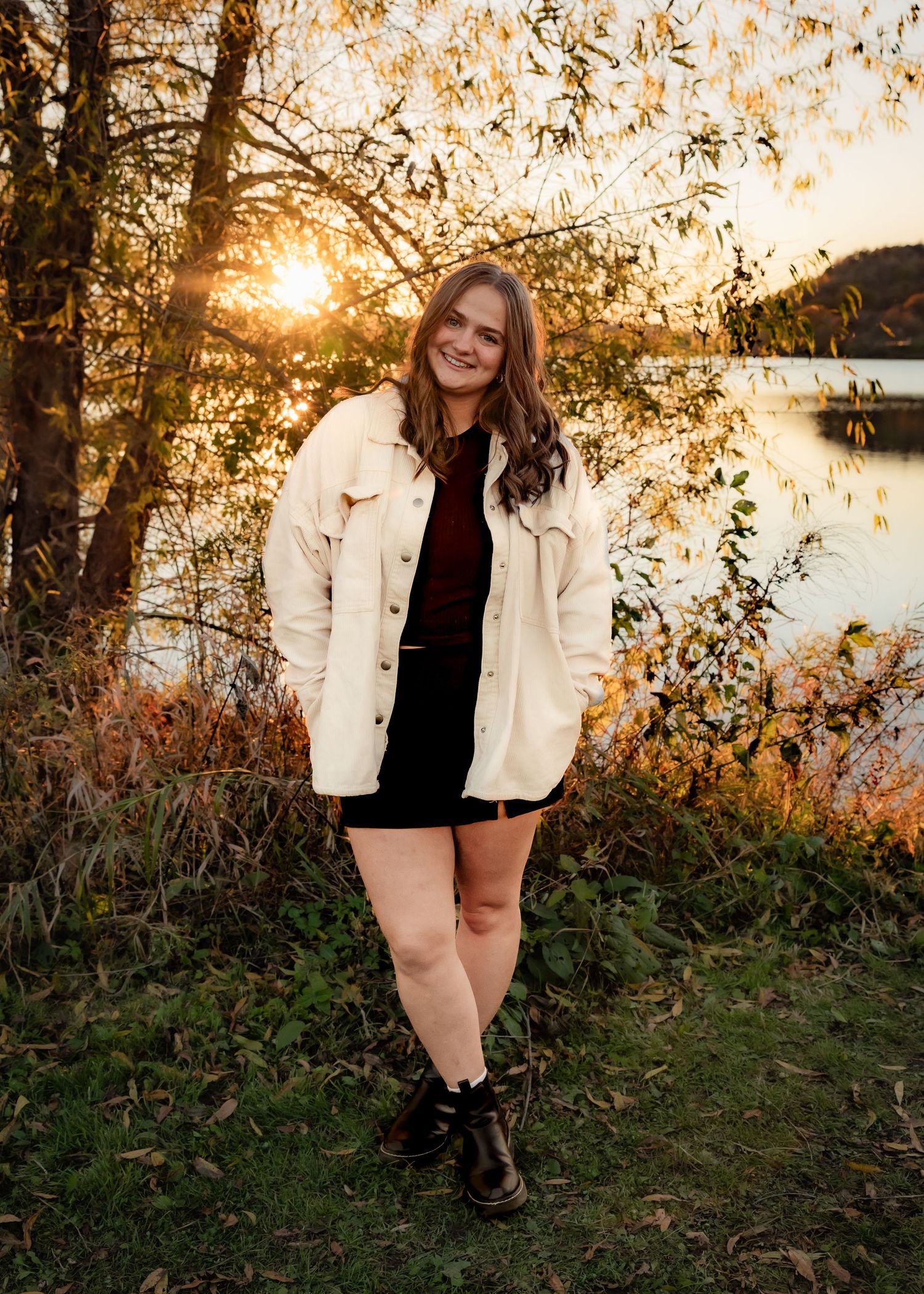 Young woman smiling in cream jacket and black skirt, posing by a lake at golden sunset surrounded by autumn foliage.