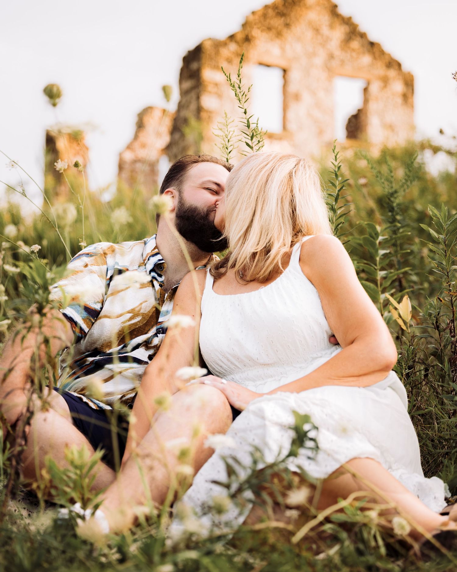 Couple sharing a romantic kiss in a wildflower field with rustic stone ruins in the background.