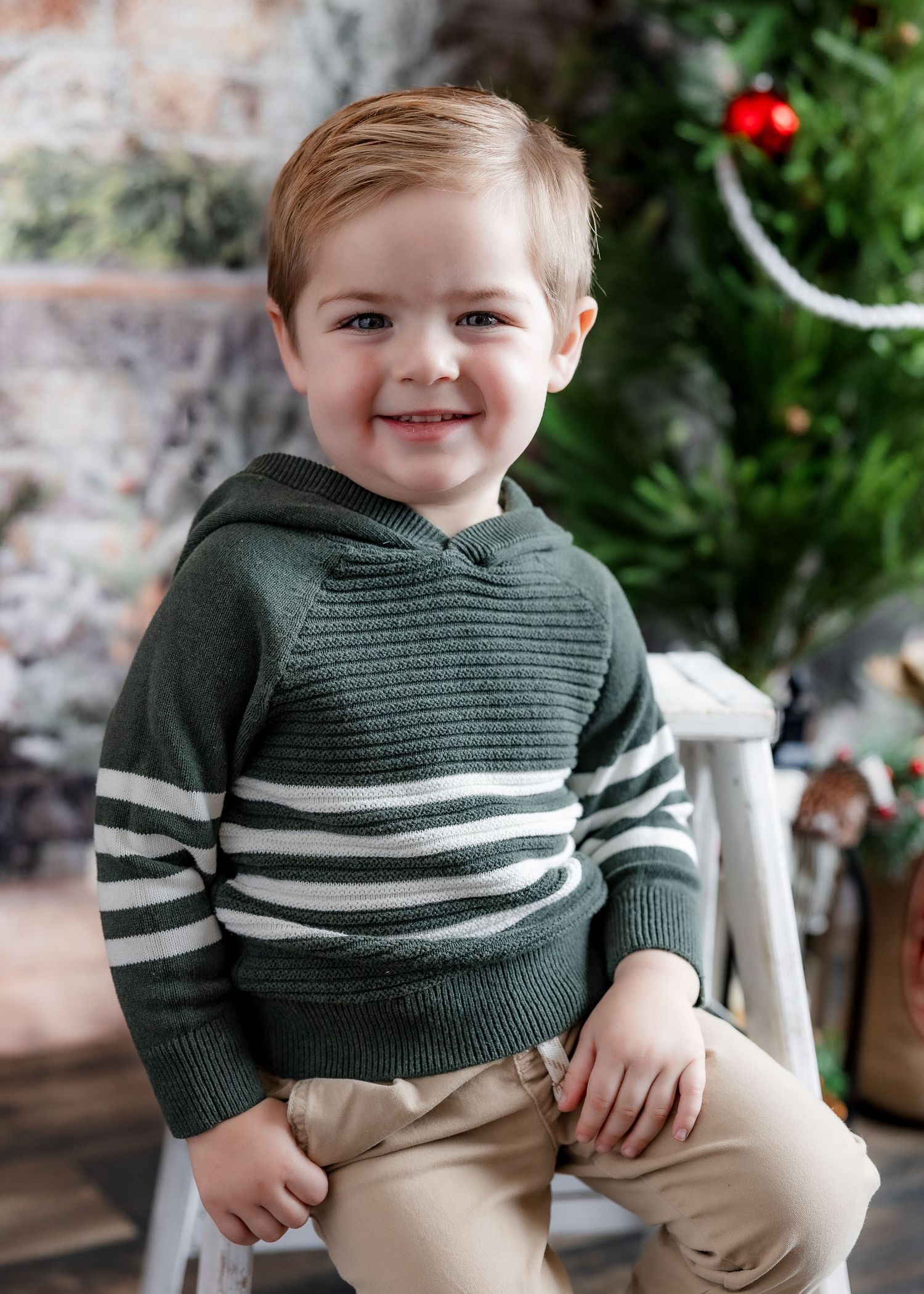 Smiling toddler boy in green striped hoodie sweater sitting near Christmas tree holiday backdrop.