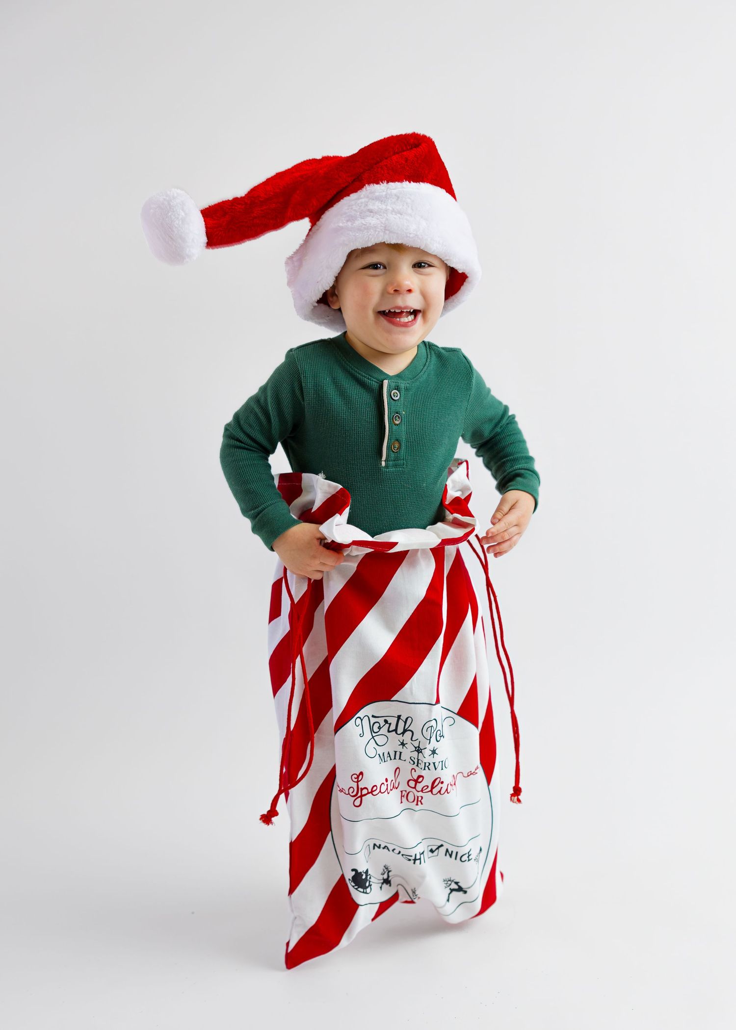 Happy toddler in green shirt and Santa hat holding a candy cane striped Christmas gift sack.