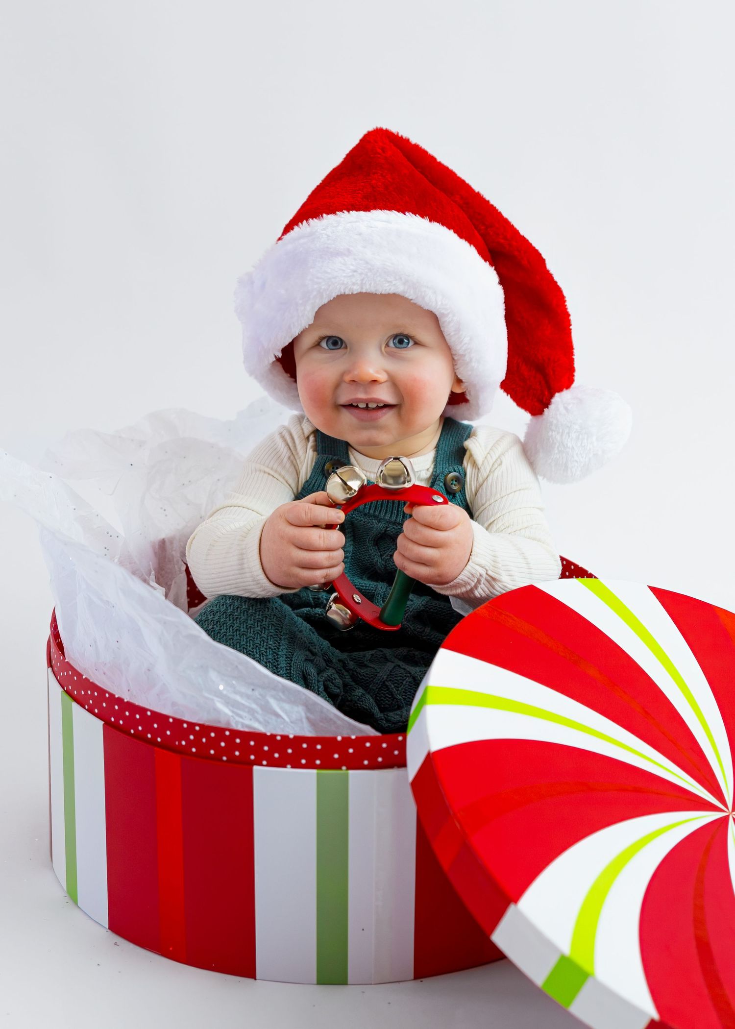 Happy baby in Santa hat sitting inside festive red striped gift box holding Christmas toy.