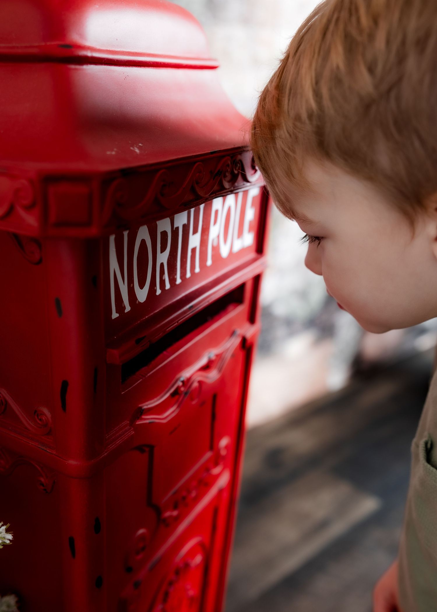 A red-haired child peeks curiously into the slot of a bright red North Pole mailbox outdoors.