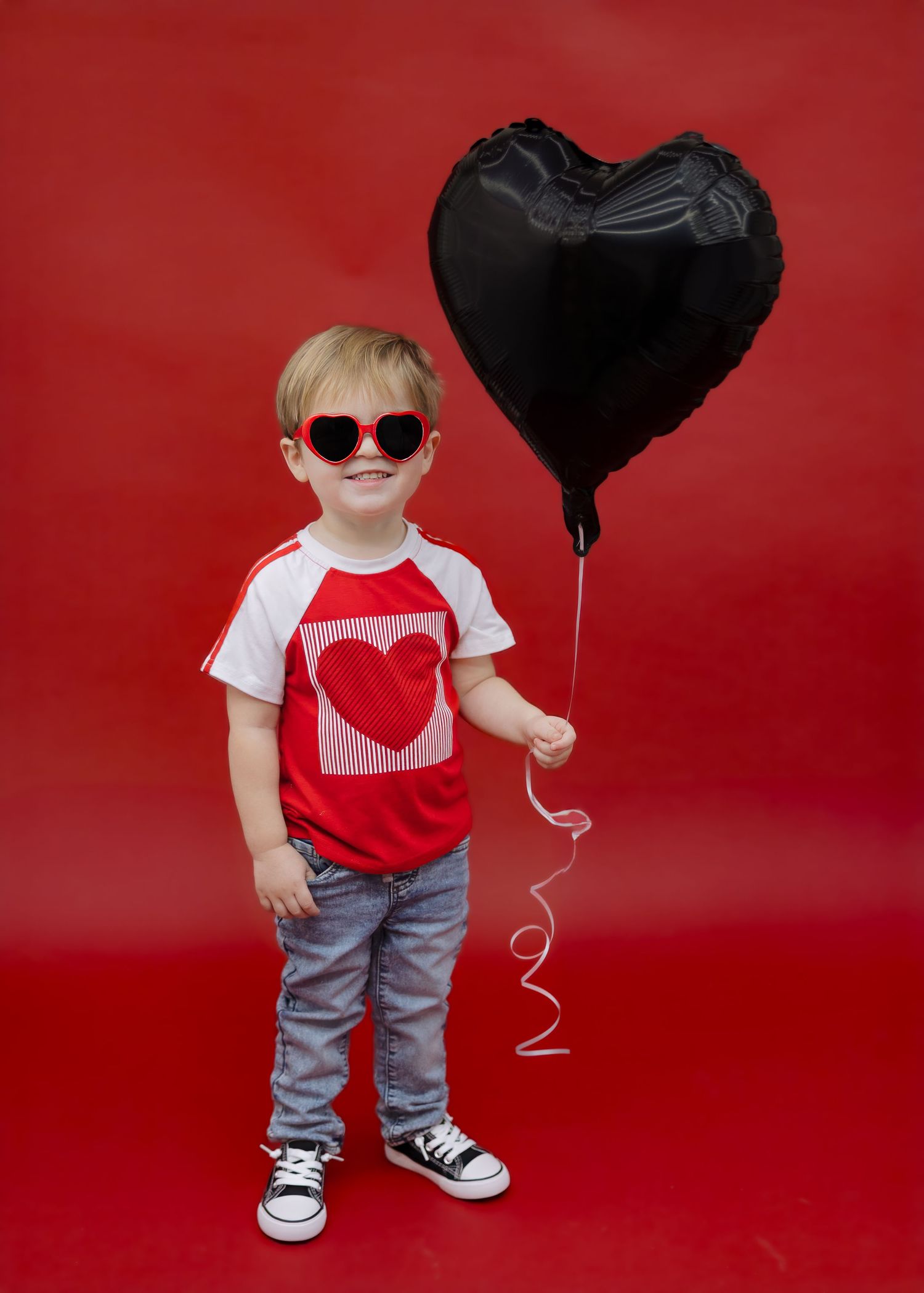 Stylish boy in heart sunglasses holding black heart balloon on vibrant red background.