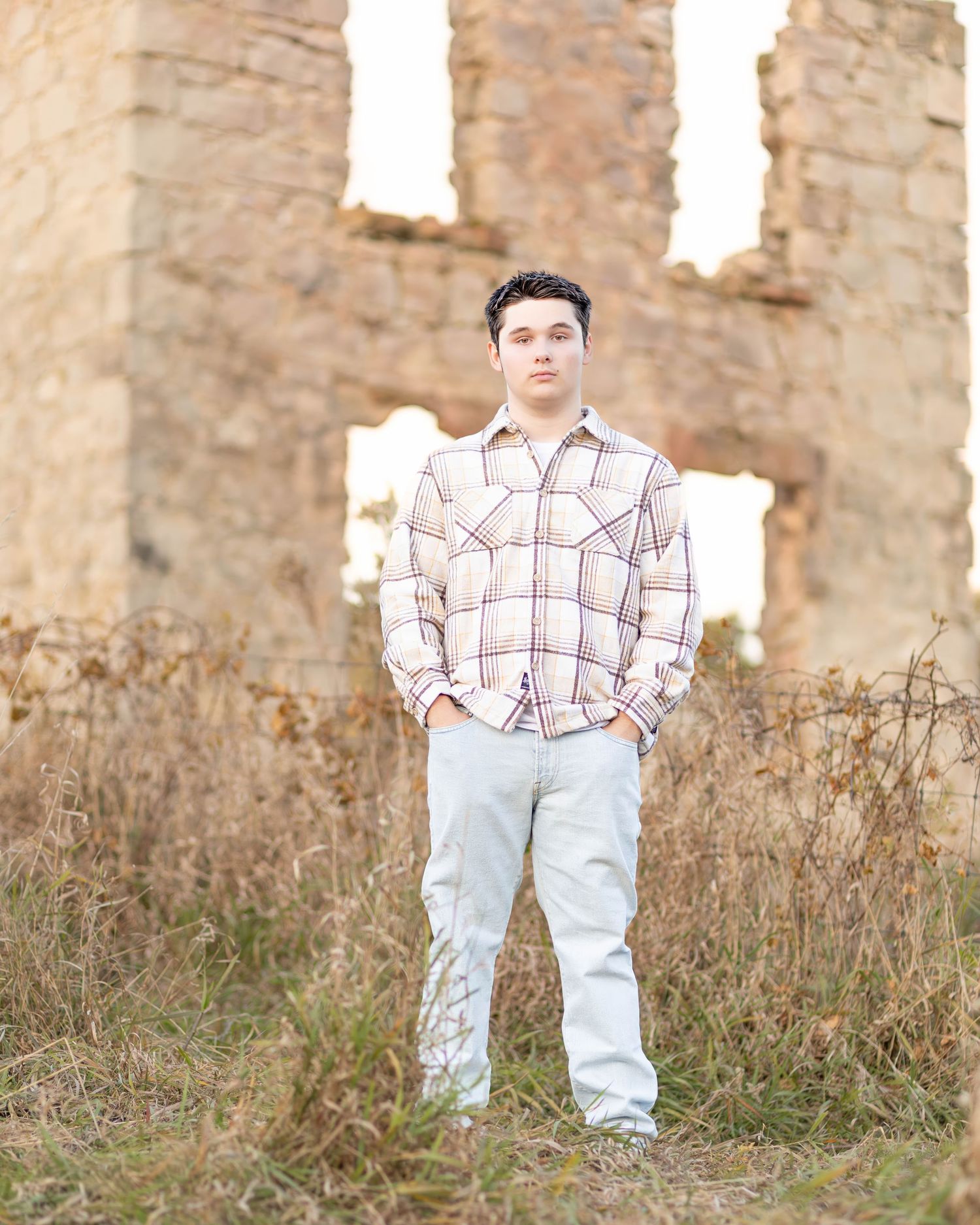 Senior boy in plaid shirt stands in tall grass before stone ruins at sunset.