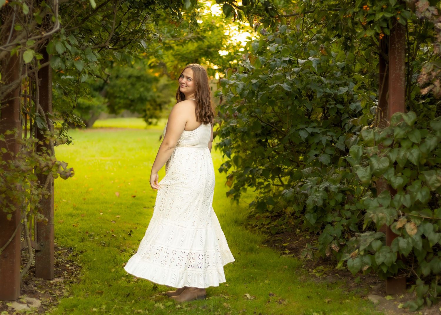 Young woman in white lace maxi dress standing between lush green trees in sunlit garden pathway.