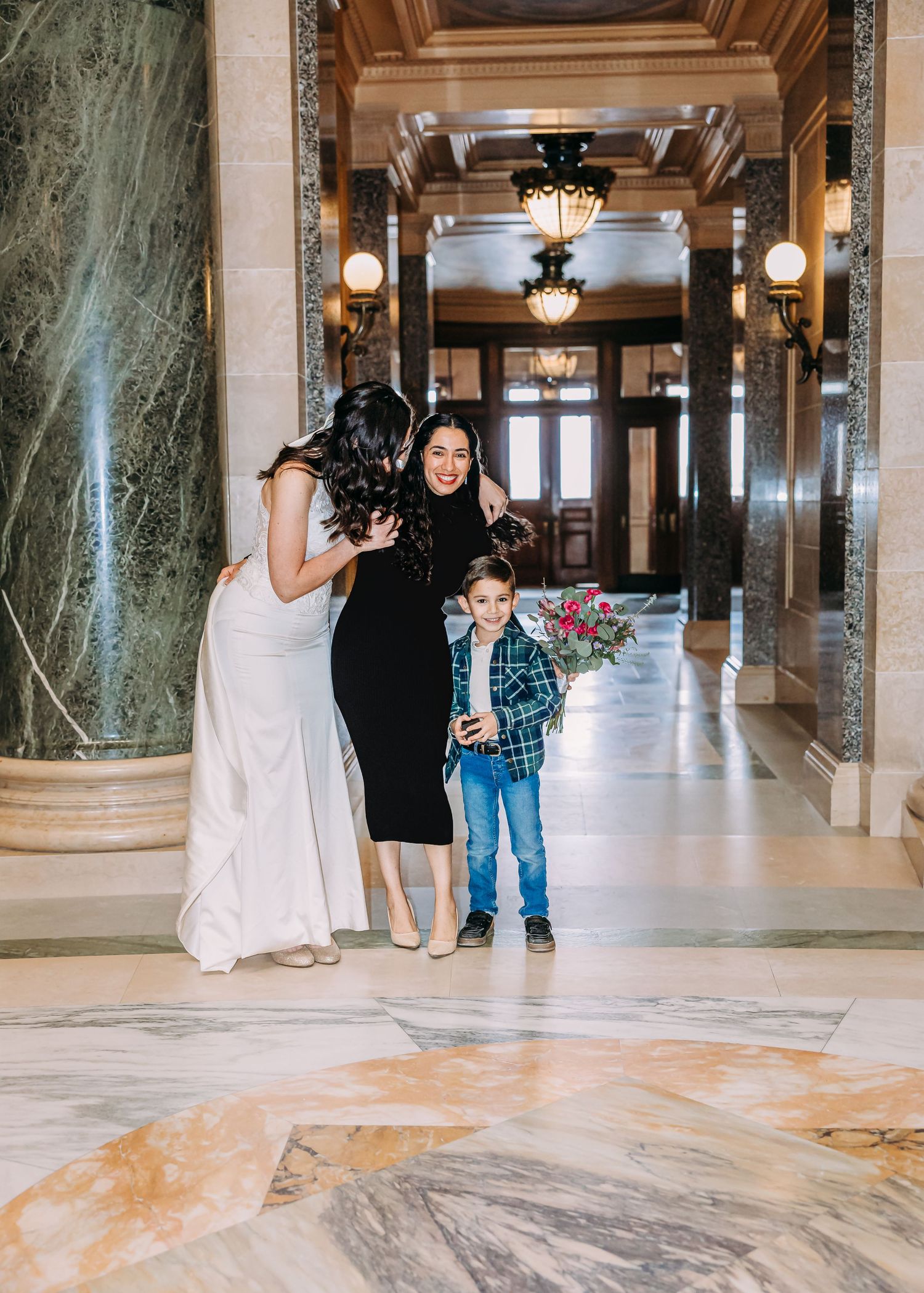 Bride in white dress embraces woman in black while young boy holds flowers in ornate marble corridor.