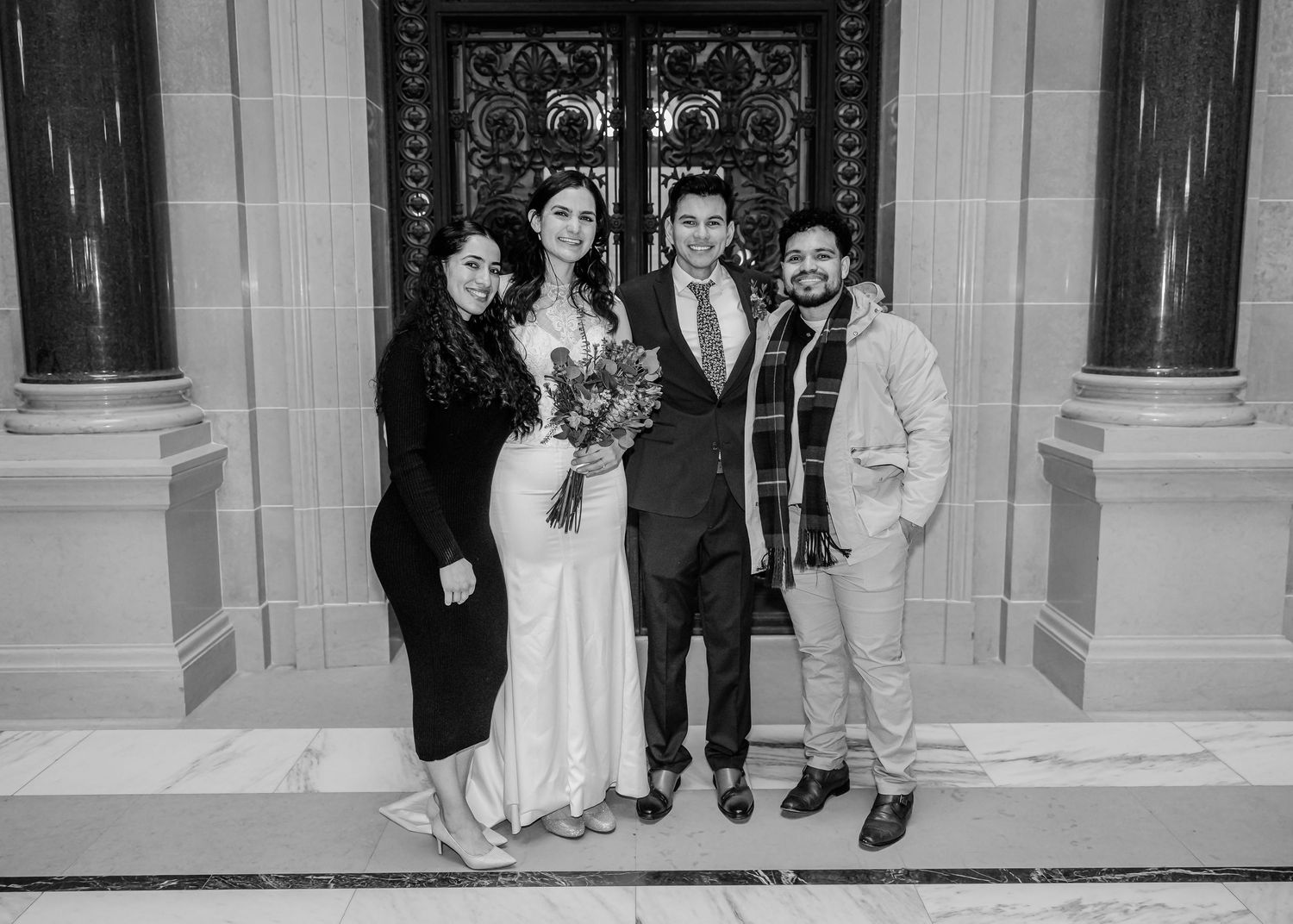 A bride and groom pose with two guests before ornate doors in a black and white wedding photo.