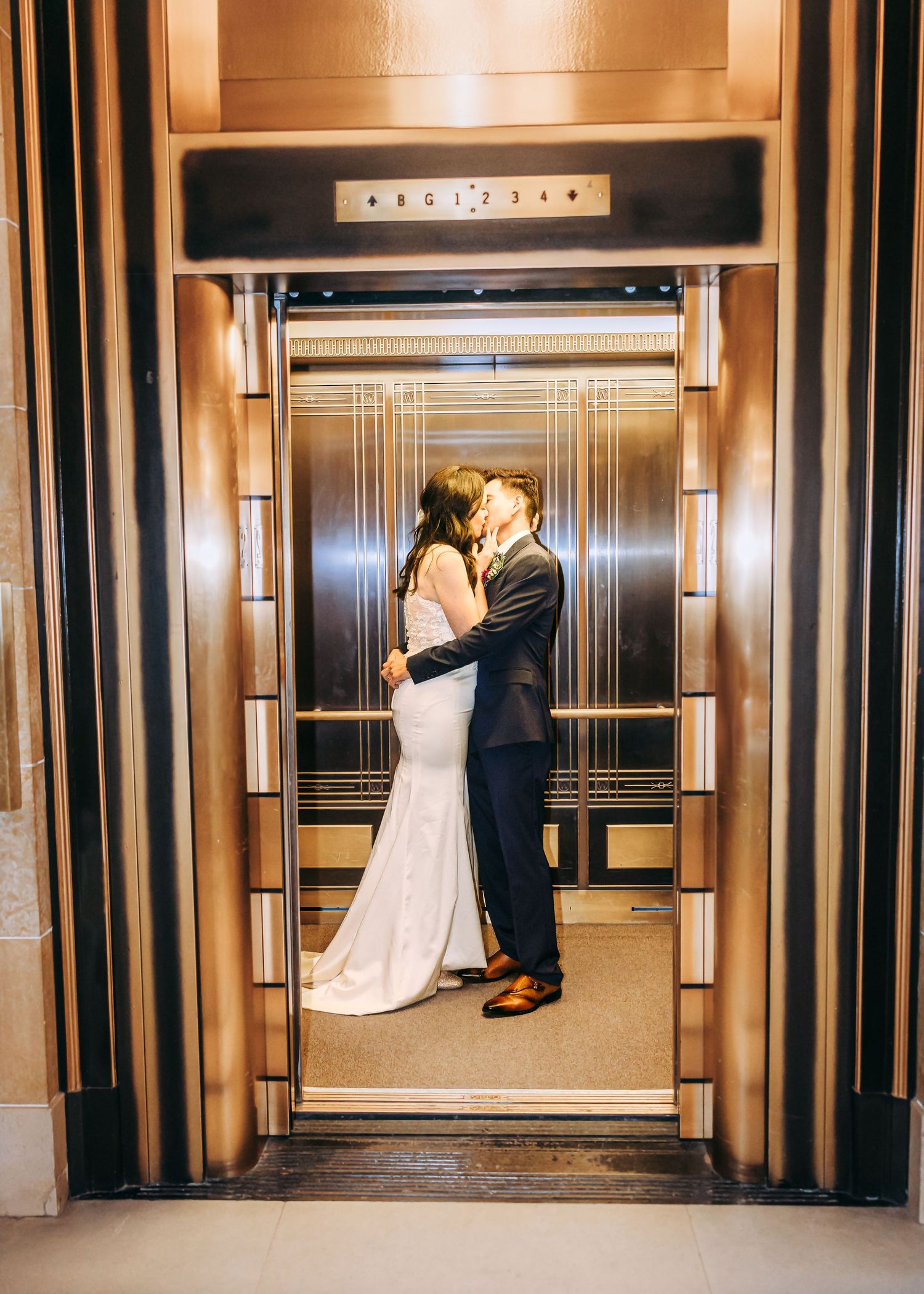 Bride and groom share a romantic kiss inside a golden Art Deco elevator with ornate details.