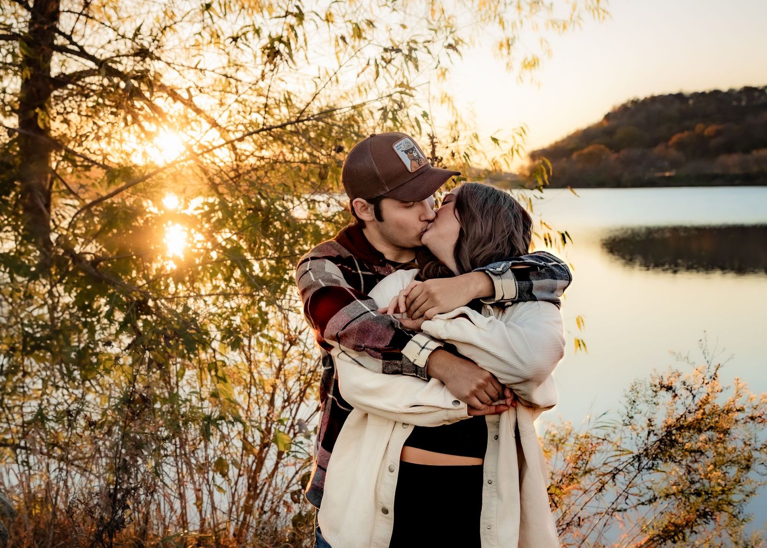 Young couple kissing by a lake surrounded by autumn foliage at golden sunset.