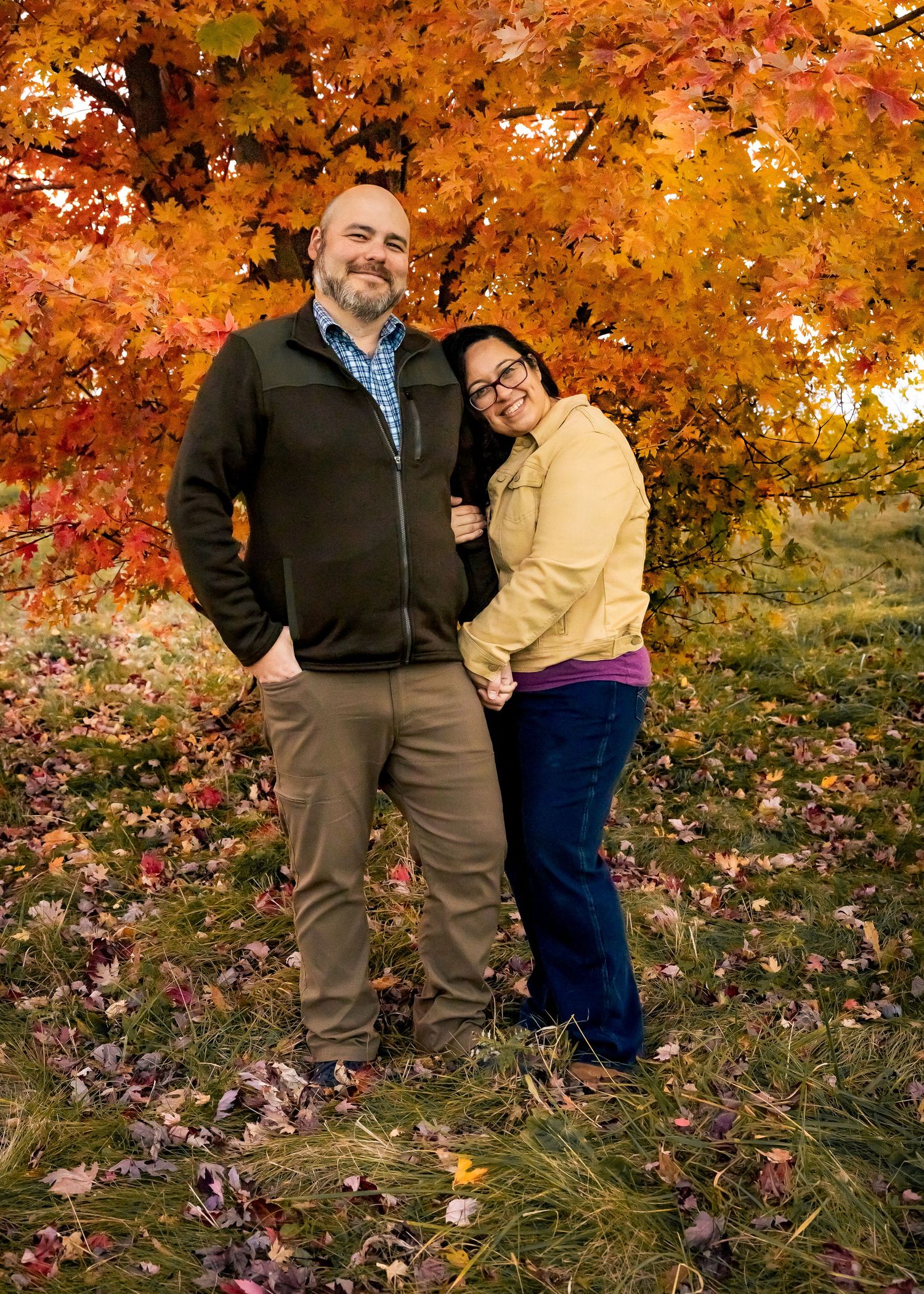 Happy couple holds hands smiling under vibrant orange and red fall foliage tree.