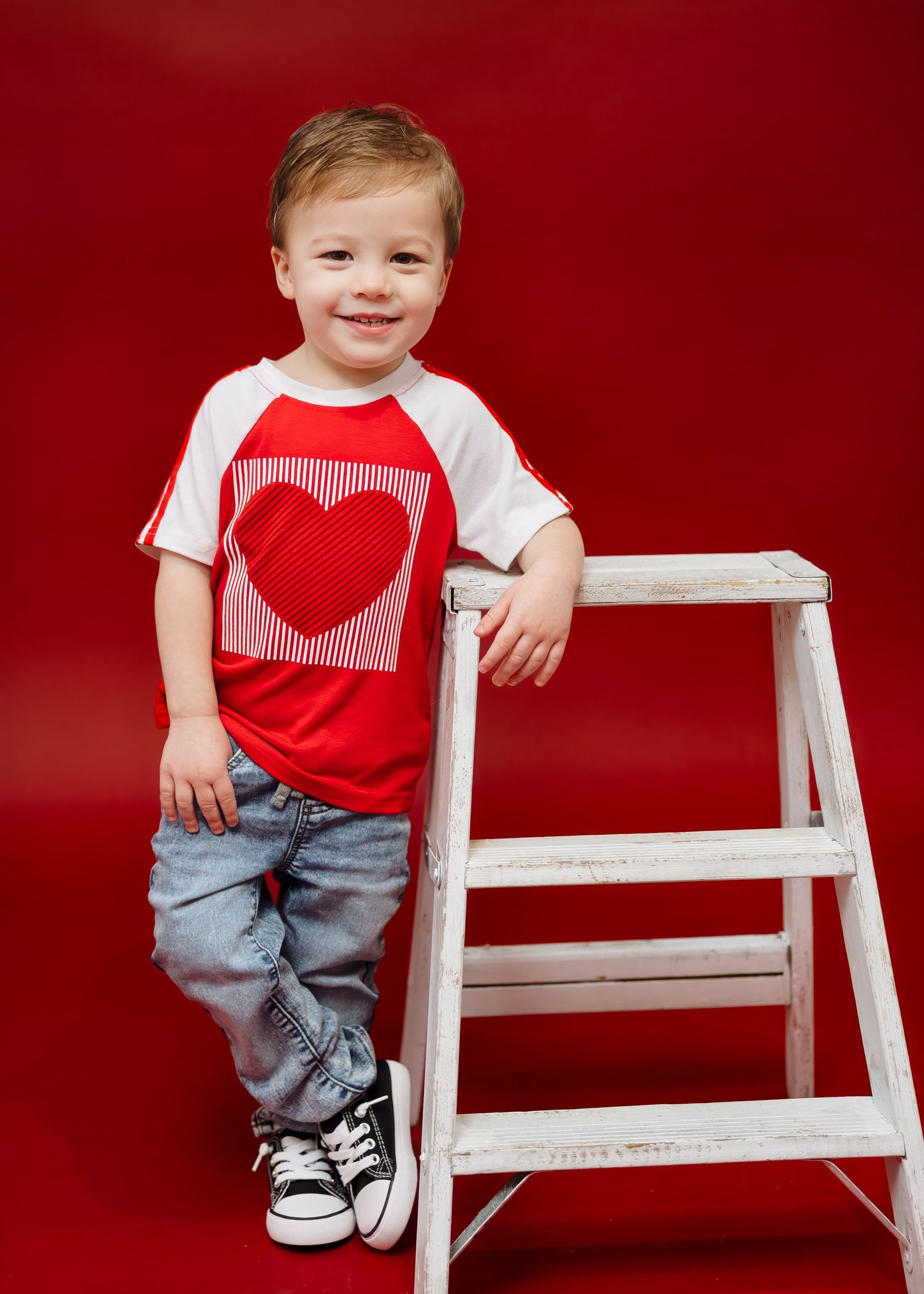 Smiling toddler in red heart shirt leans on white ladder, red backdrop.