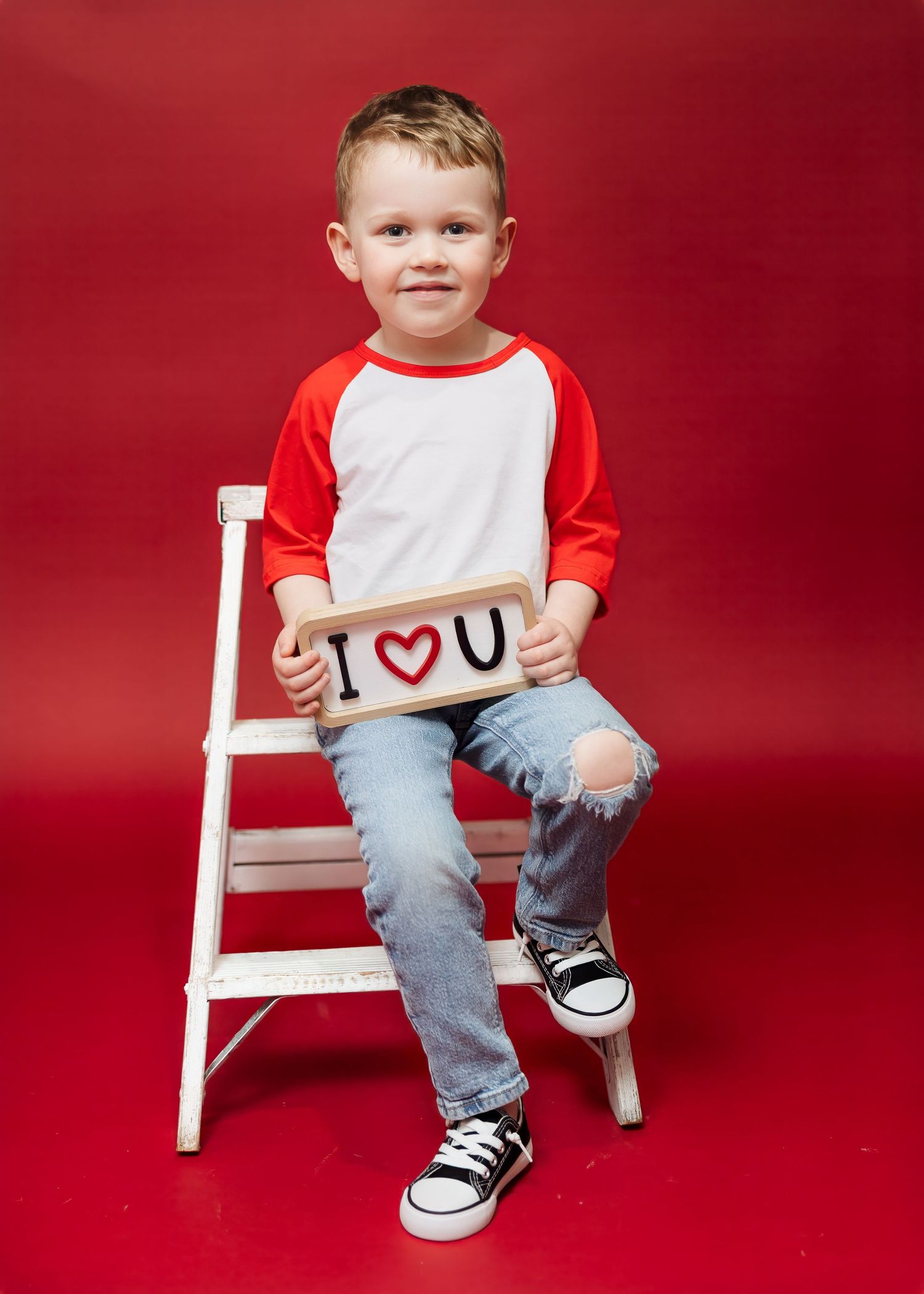 Young boy sits on white ladder holding I Love You sign on red background.