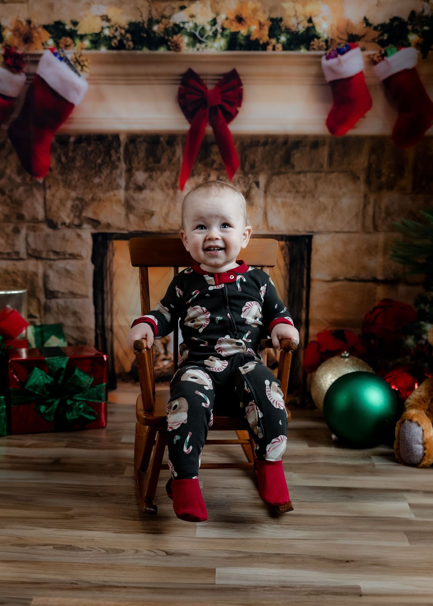 Smiling baby in black Santa pajamas sitting in wooden rocking chair by Christmas fireplace.
