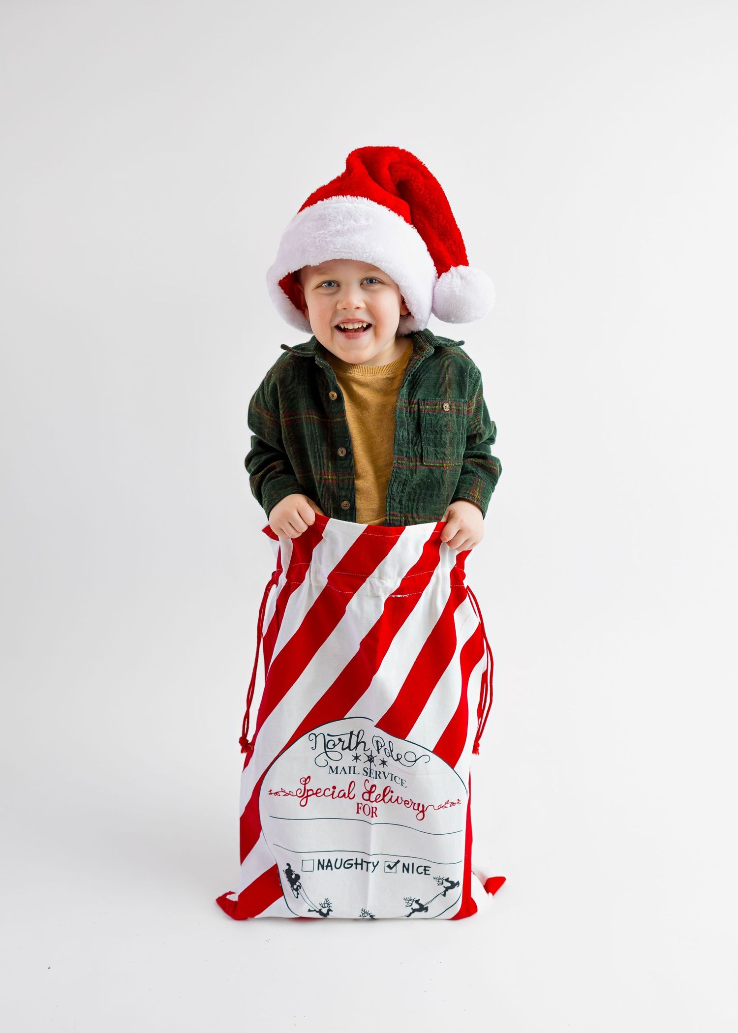 Smiling toddler in green shirt and Santa hat stands inside a red striped Christmas sack.