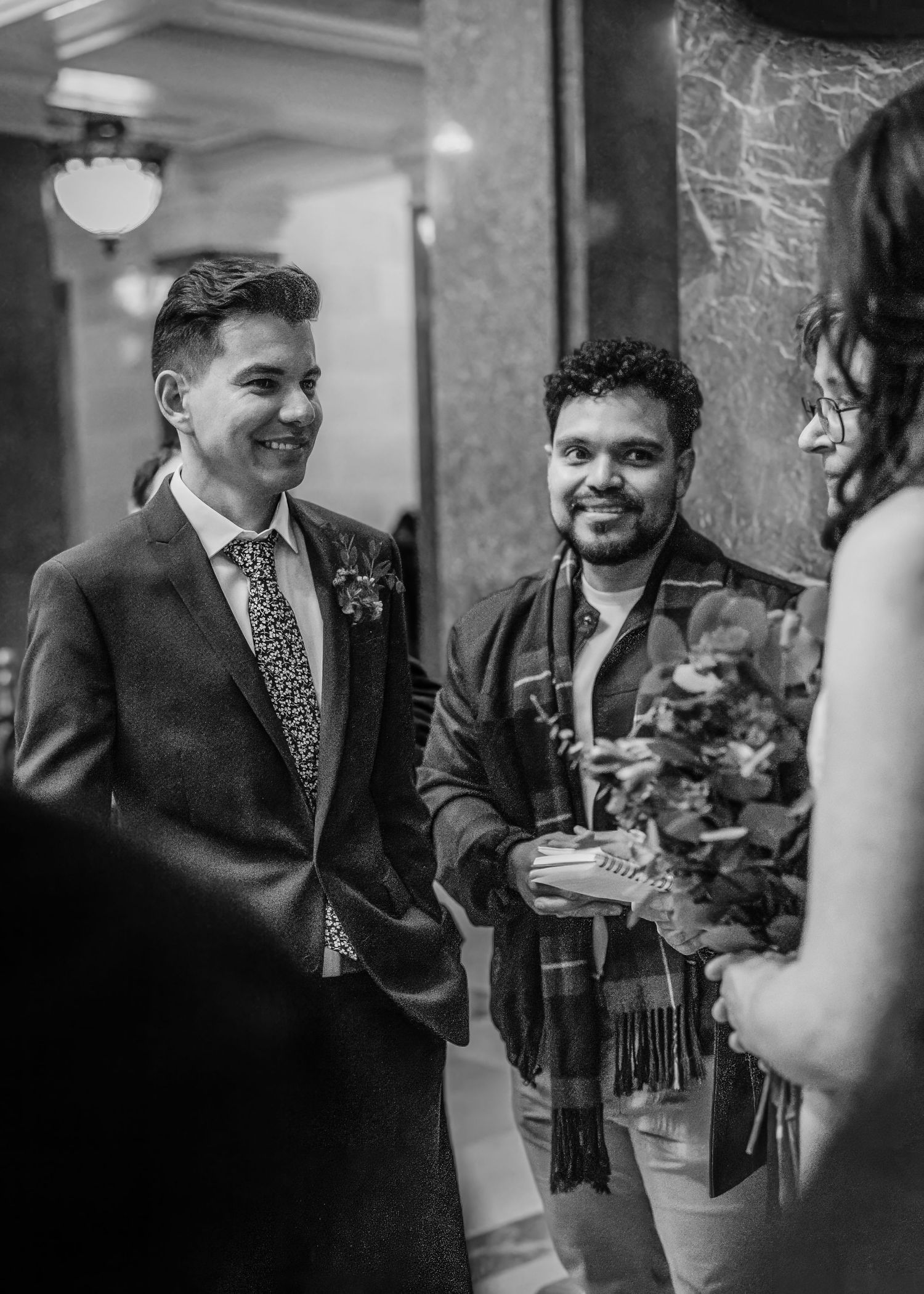 Groom in navy suit smiles with guest holding bouquet during wedding ceremony in elegant venue.