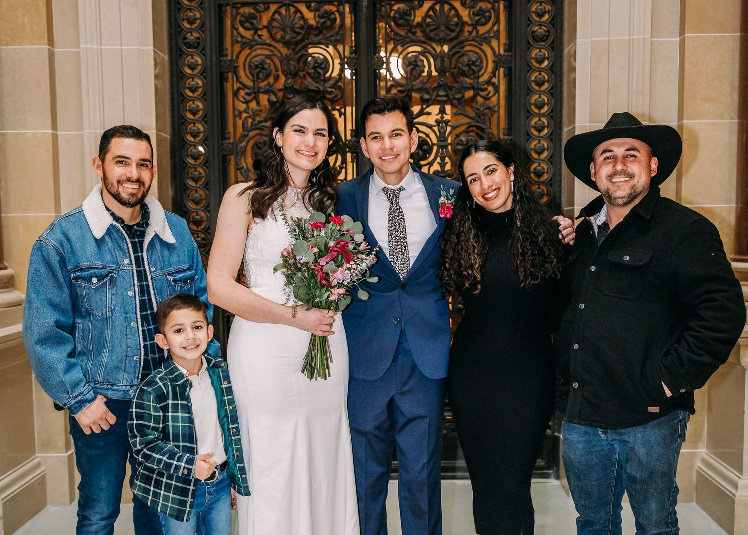Bride and groom celebrate with five family members including a young child in front of ornate golden doors.