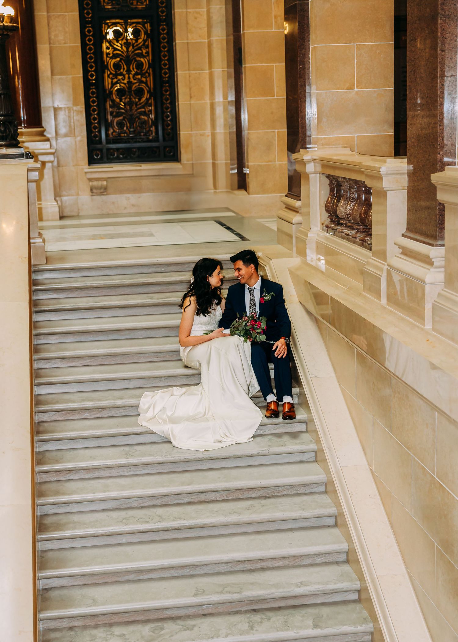 Bride in white gown and groom in navy suit sitting together on marble stairs in a grand historic building.