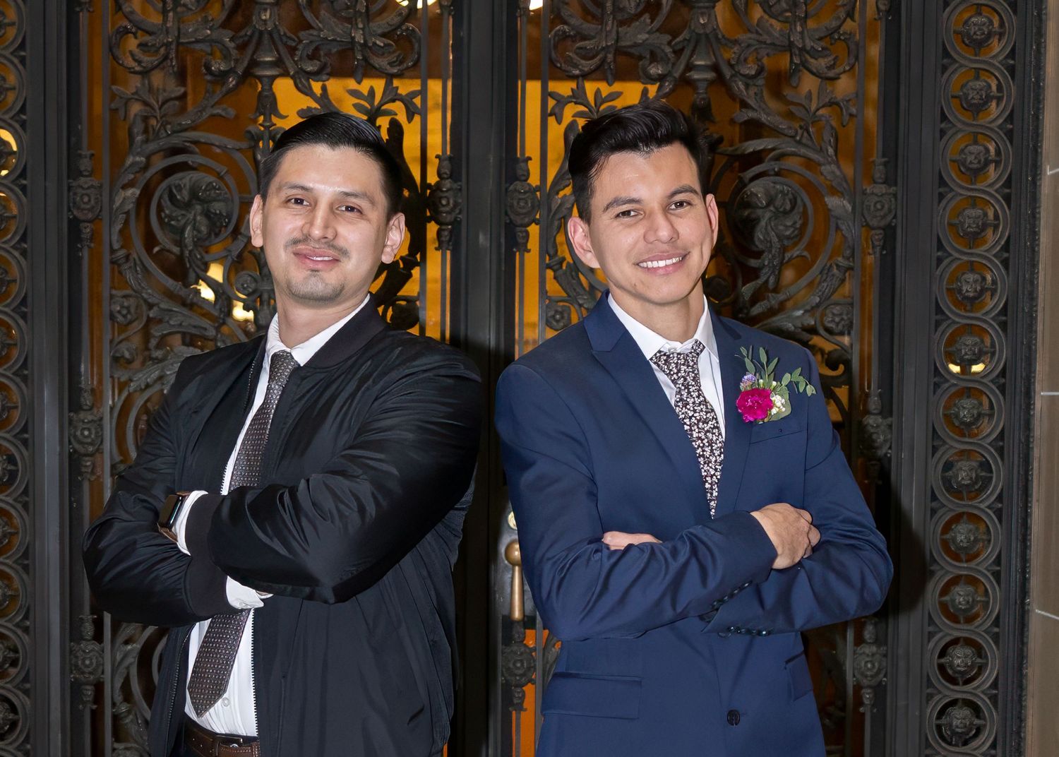 Two well-dressed men in suits posing back-to-back before ornate golden doors at a wedding.