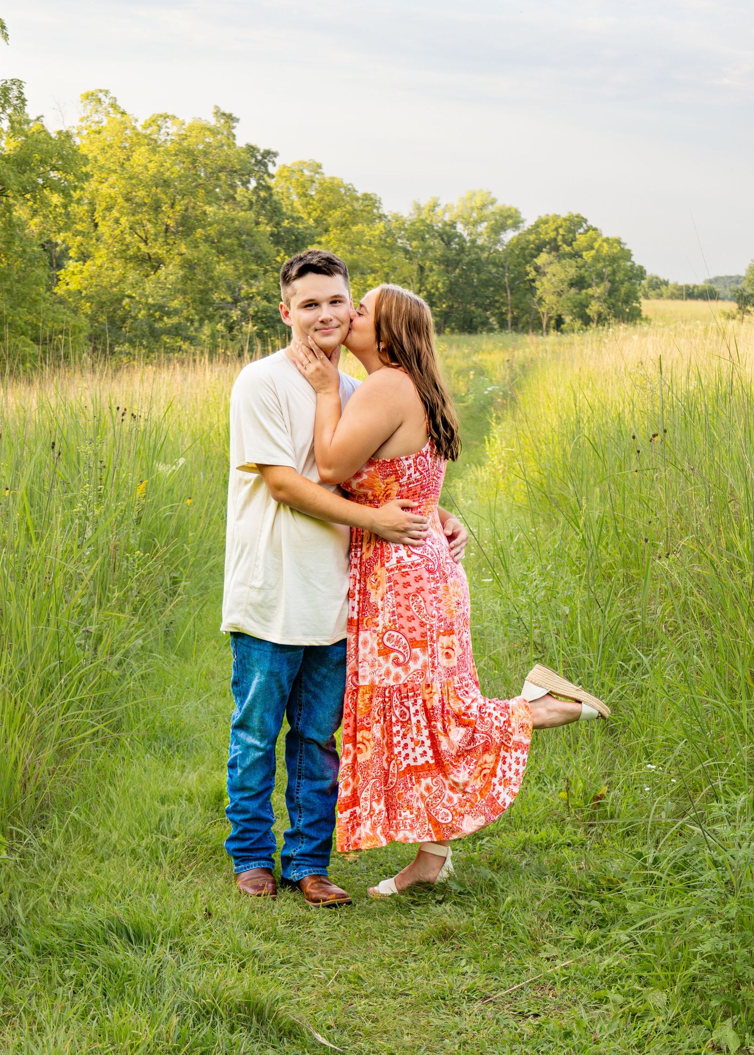 Young woman in pink floral maxi dress kissing mans cheek while he smiles in green field setting.