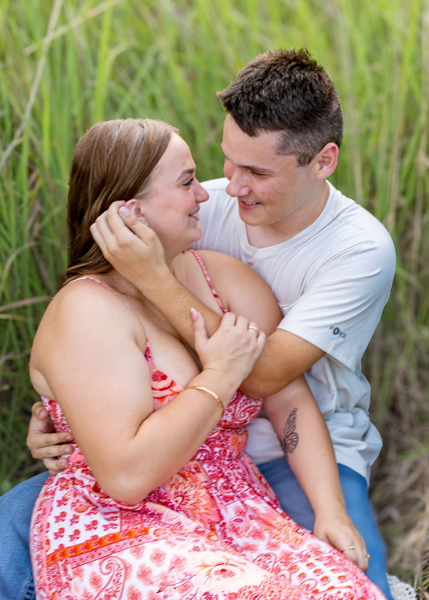 Couple sitting closely in tall green grass, sharing a joyful romantic outdoor moment.
