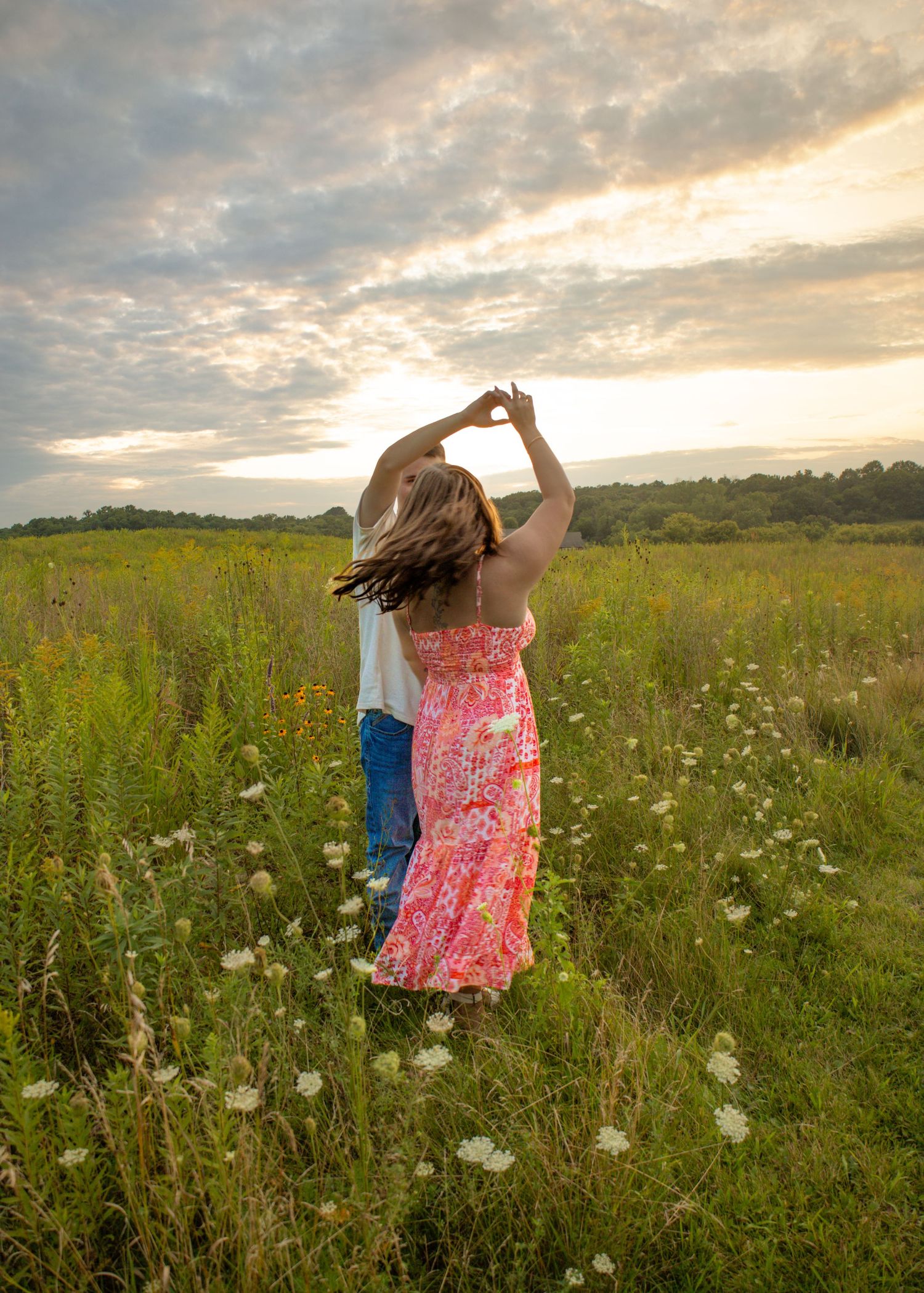 Romantic couple dancing together from behind in a lush green wildflower field during a golden sunset.