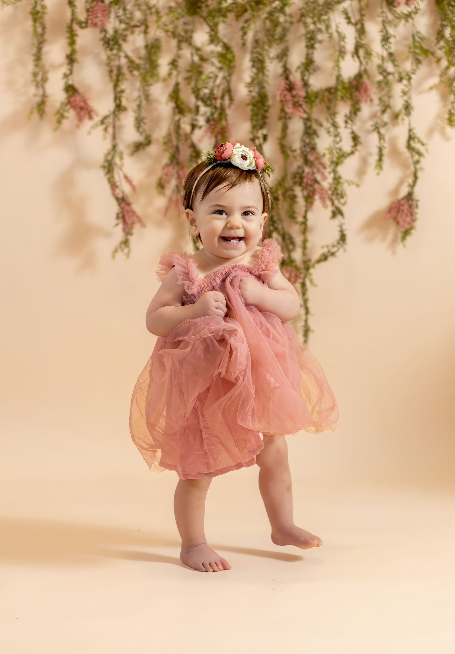 Happy baby girl in a pink tulle dress stands smiling beneath hanging floral greenery.