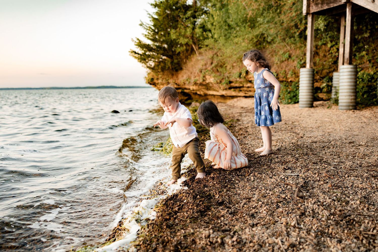 Three children playing on a rocky beach shore at sunset, exploring the water's edge near a wooded coastline.