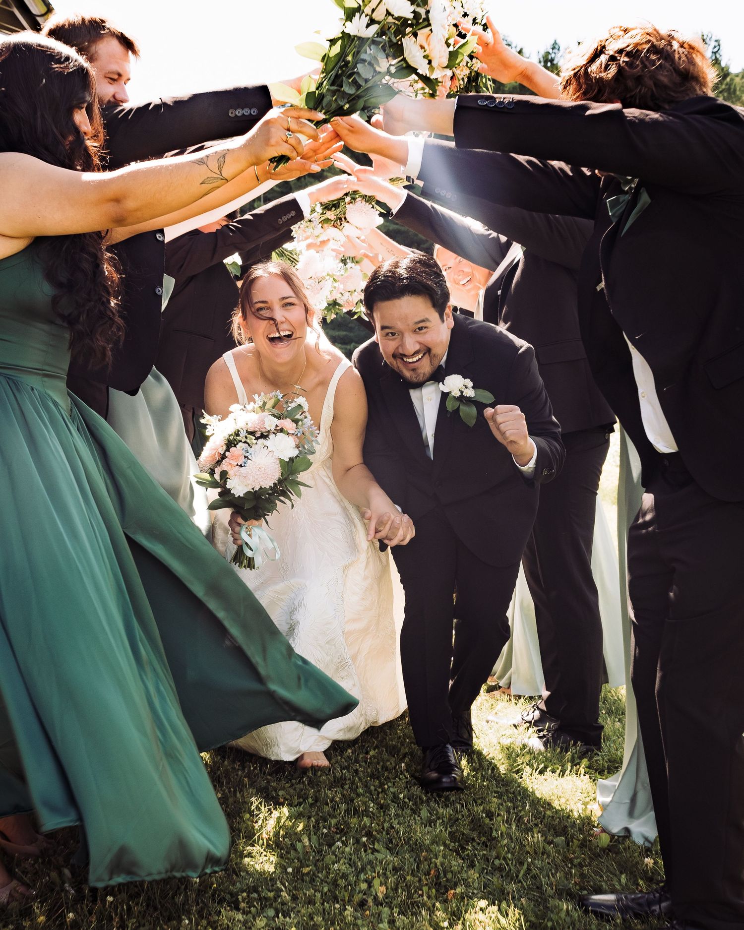 Joyful bride and groom run through a tunnel of raised hands formed by wedding party in green and black attire.