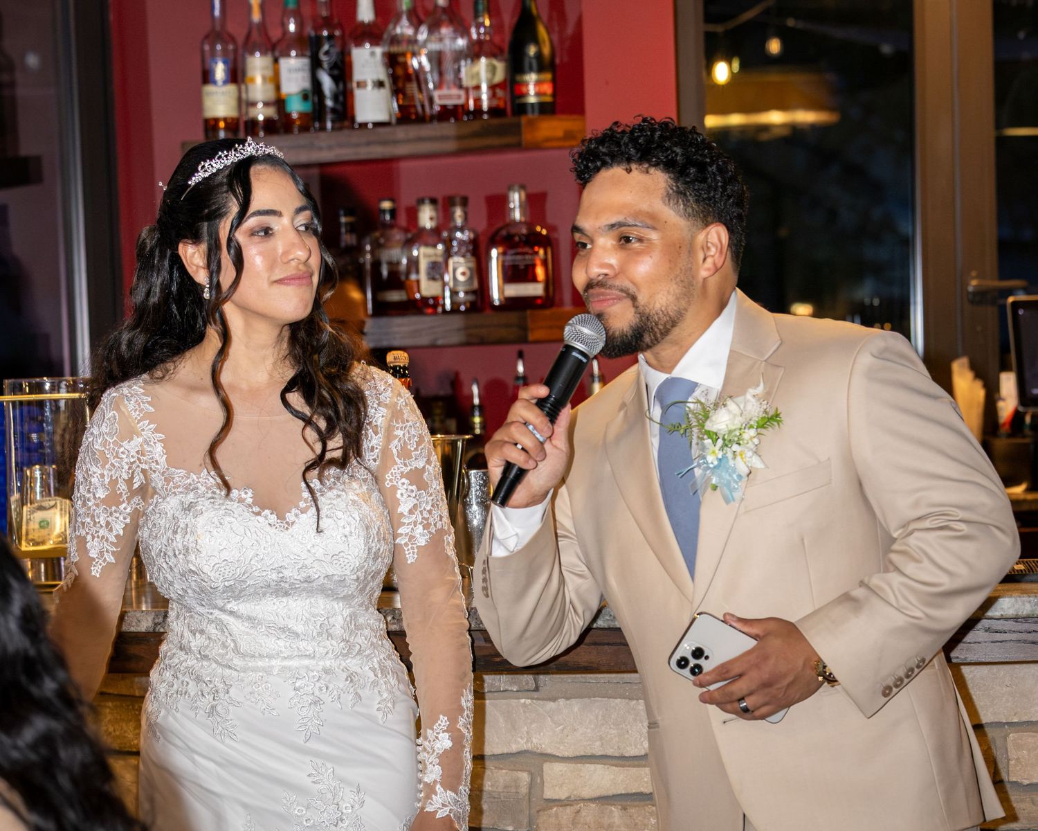 Groom in a tan suit with a boutonniere speaks into a microphone beside a smiling bride in a lace gown with a tiara at a wedding reception.