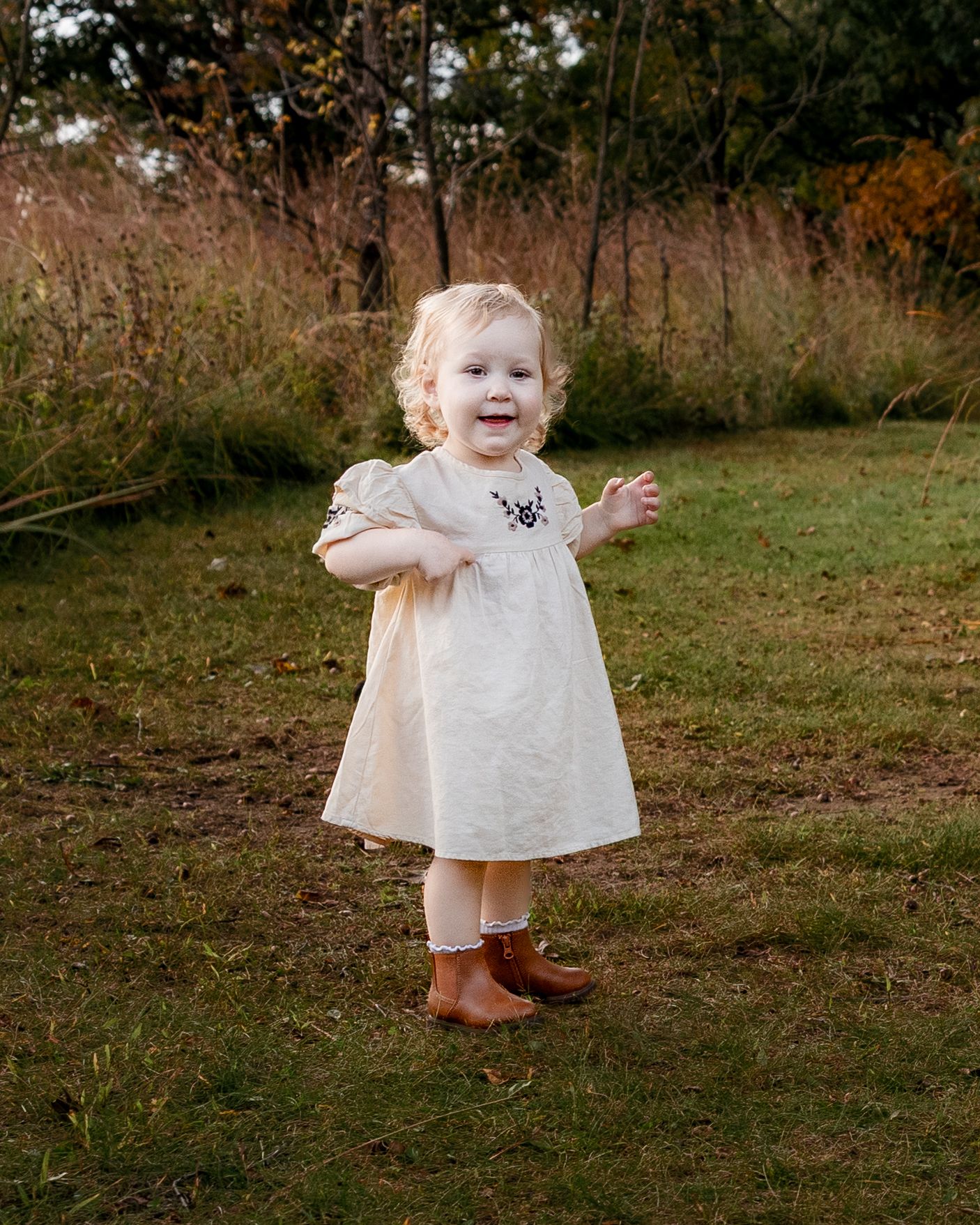 Smiling toddler girl in cream dress and brown boots standing on green grass in an autumn outdoor setting.