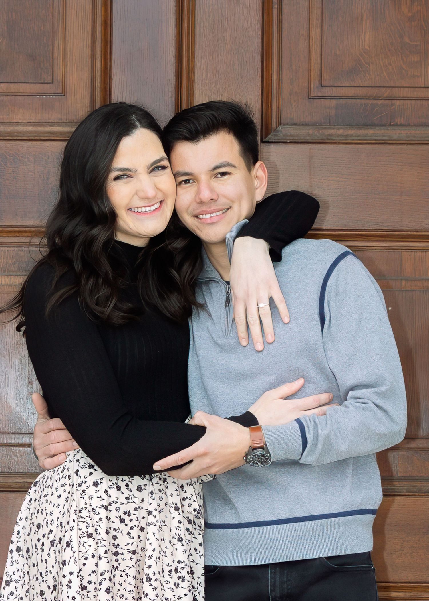 A smiling couple poses closely together in front of elegant wooden doors, the woman showing an engagement ring.