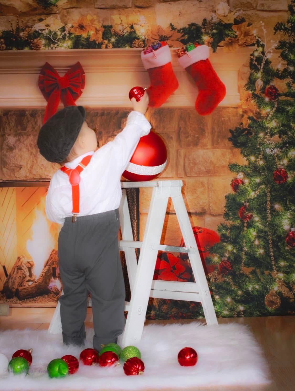 Young boy in suspenders reaches for Christmas ornament on ladder near decorated fireplace.