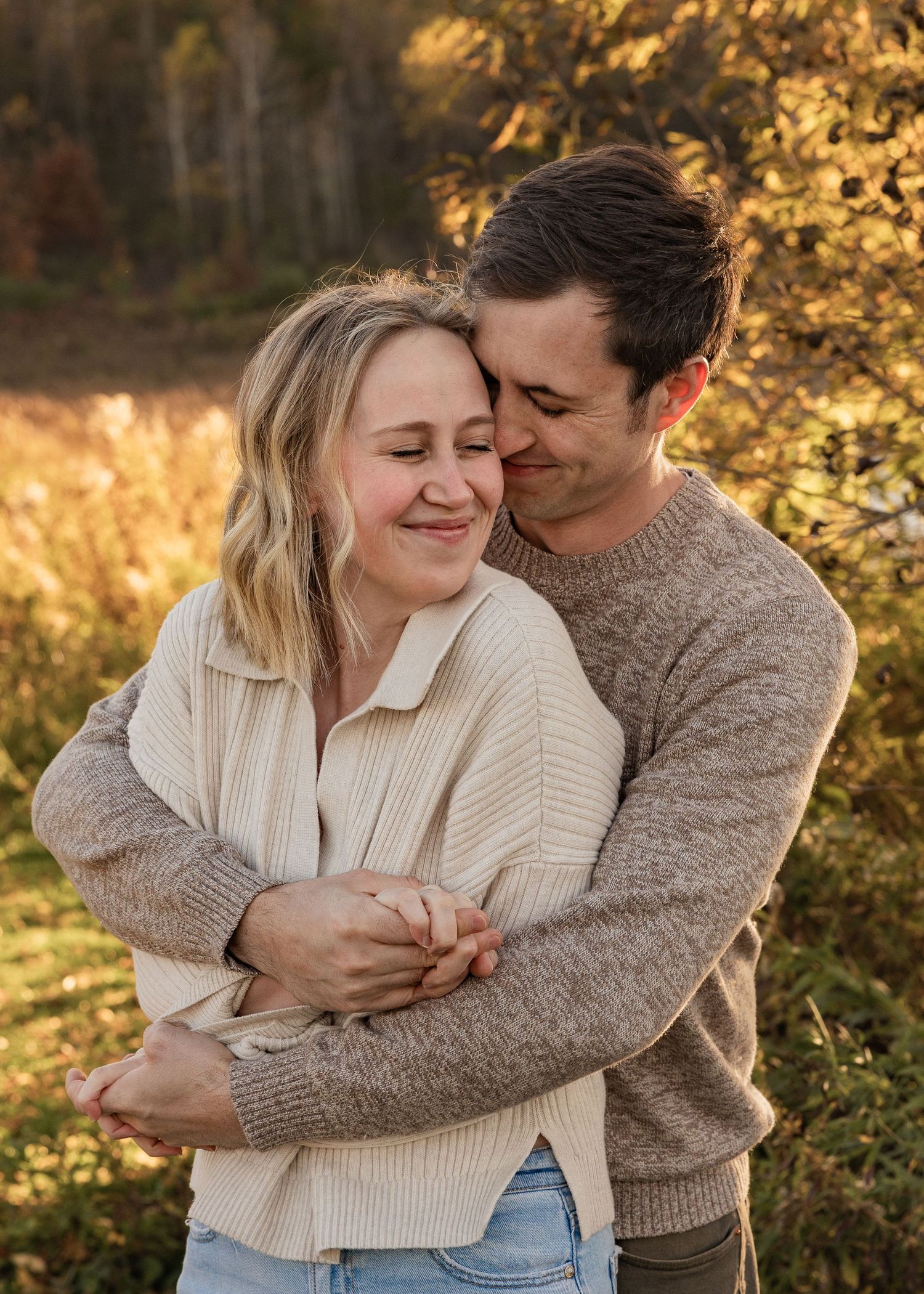 Man hugging woman from behind as she smiles with eyes closed in autumn setting.