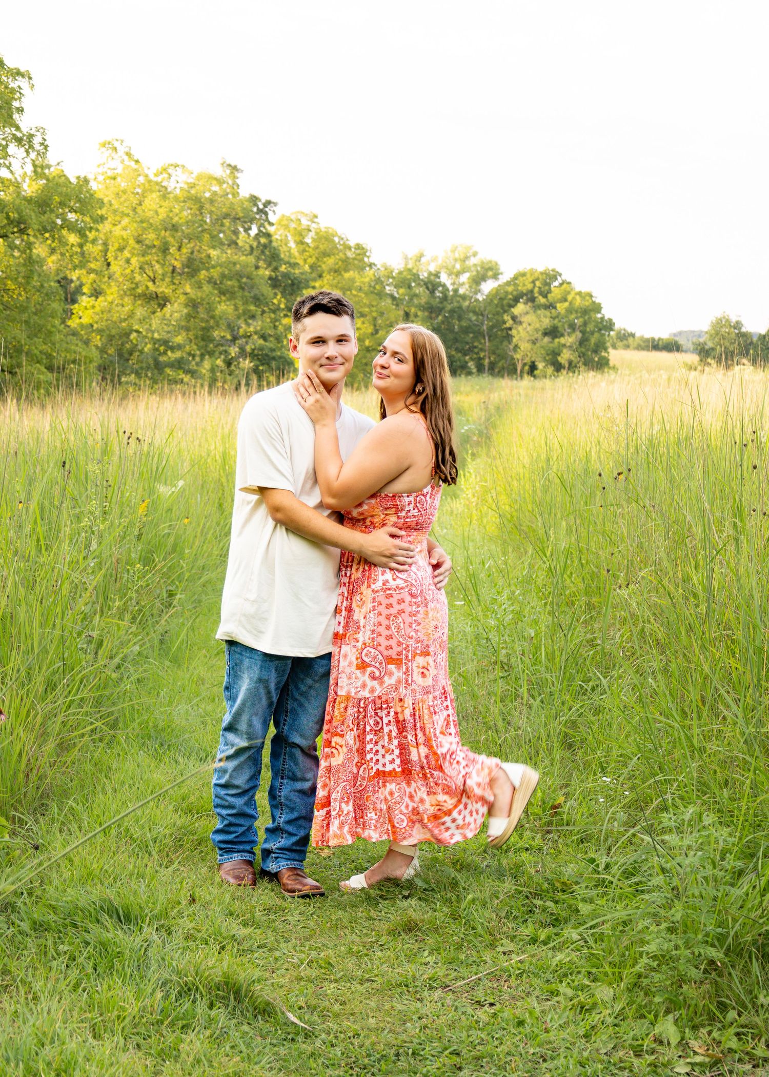 Young couple embracing on a green meadow path, woman in a coral floral maxi dress.