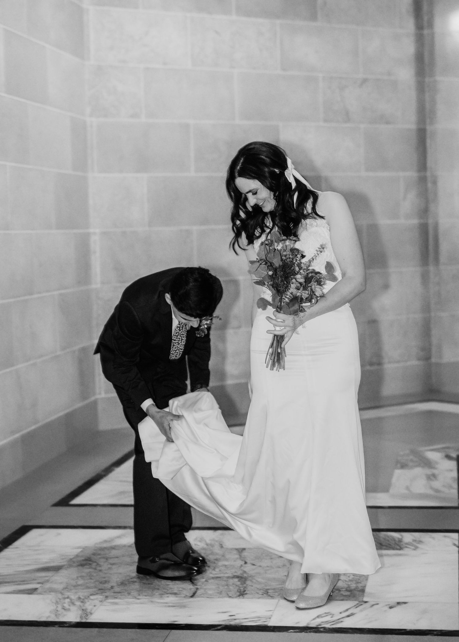 Black and white photo of groom adjusting bride's dress train as she smiles holding bouquet.