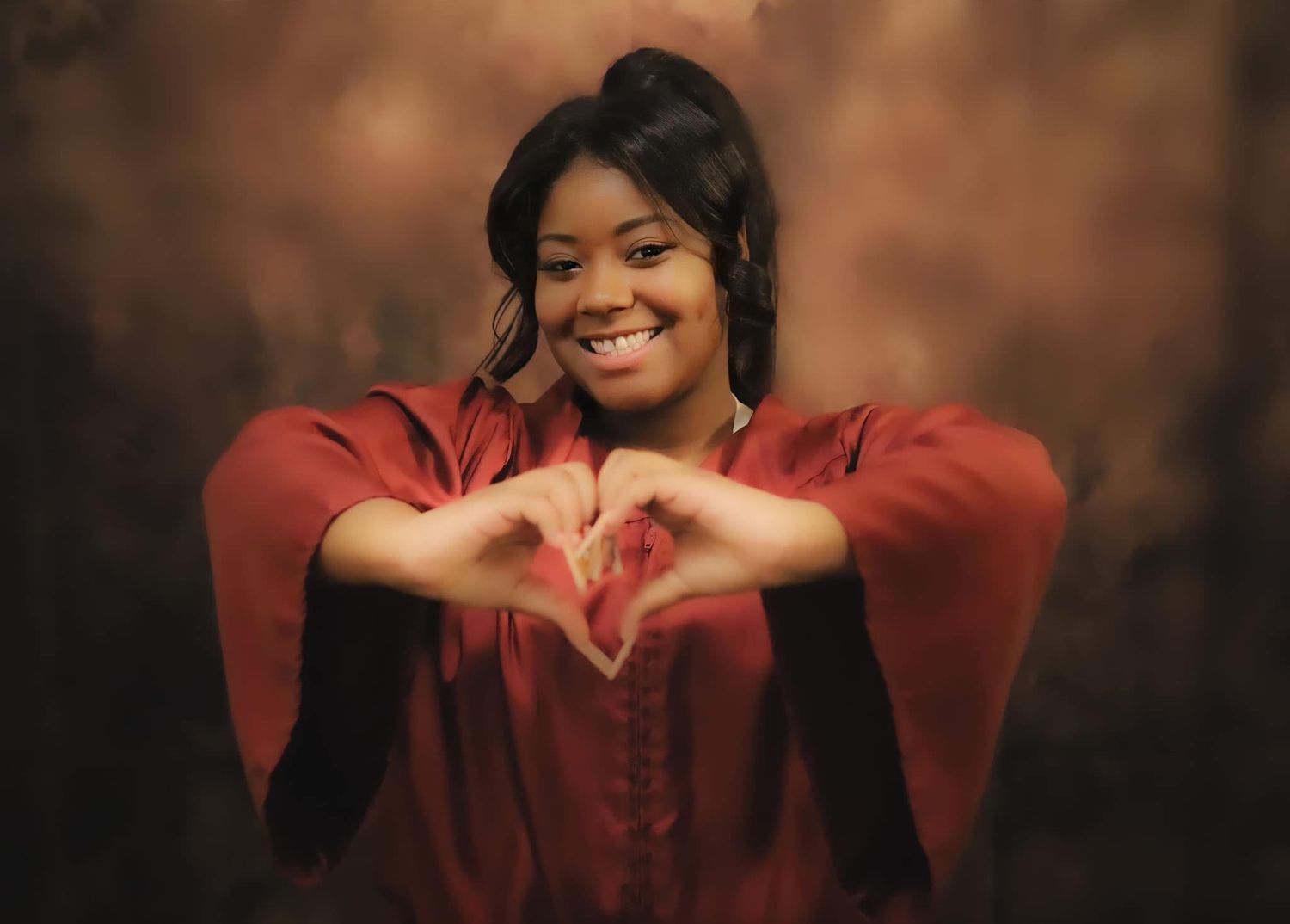 Young woman in red dress smiling and making a heart shape with her hands against a brown backdrop.