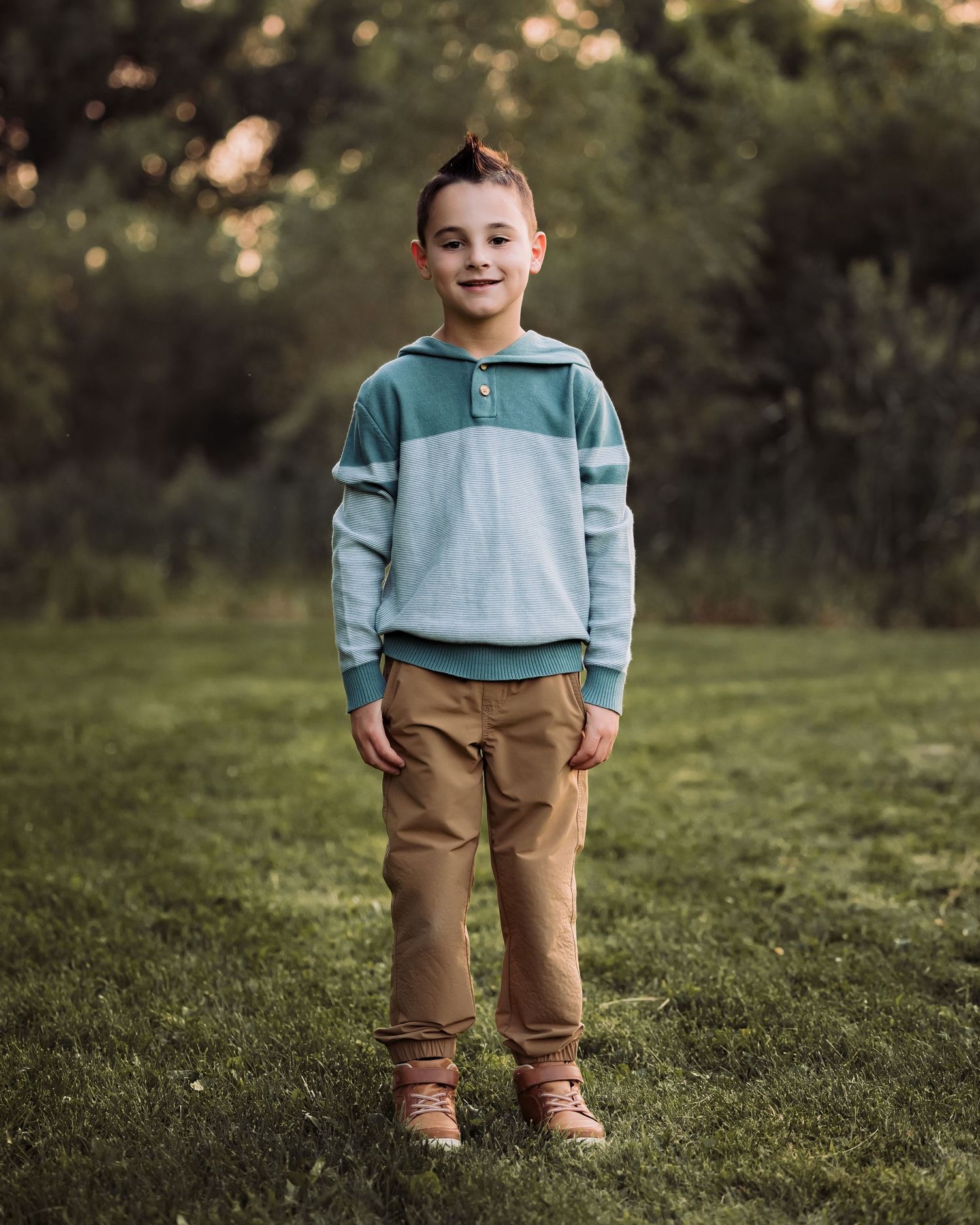 A smiling young boy stands on green grass wearing a teal sweater and tan pants in a softly lit outdoor portrait.