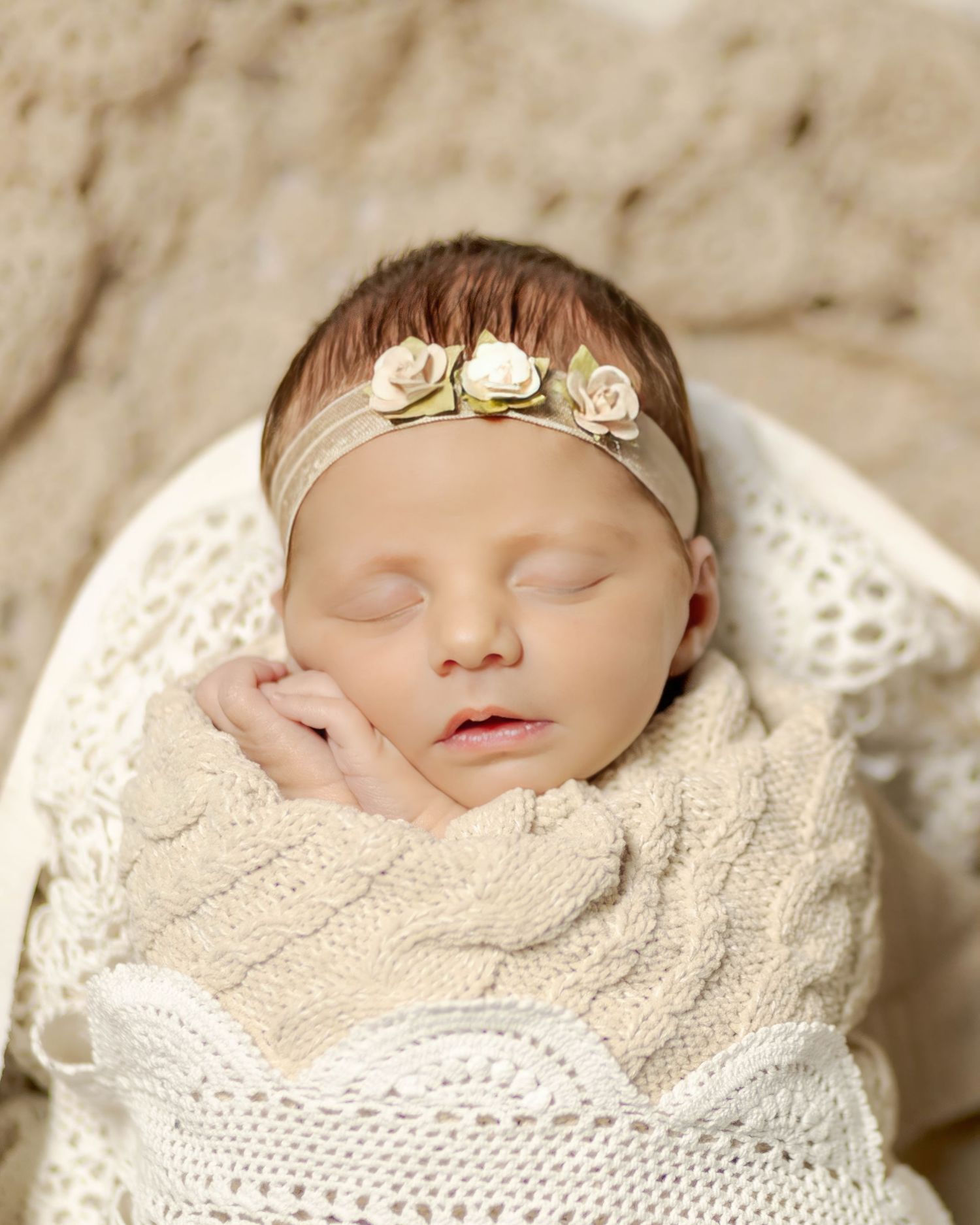 Sleeping newborn baby swaddled in cream knit blanket wearing a delicate floral headband.