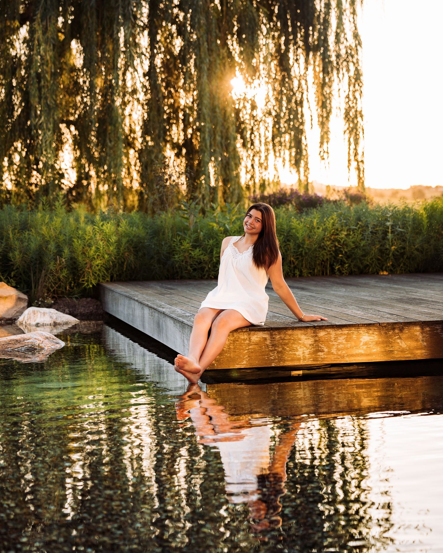 Woman in white dress sitting on a wooden dock at sunset, with willow trees and calm reflective water.