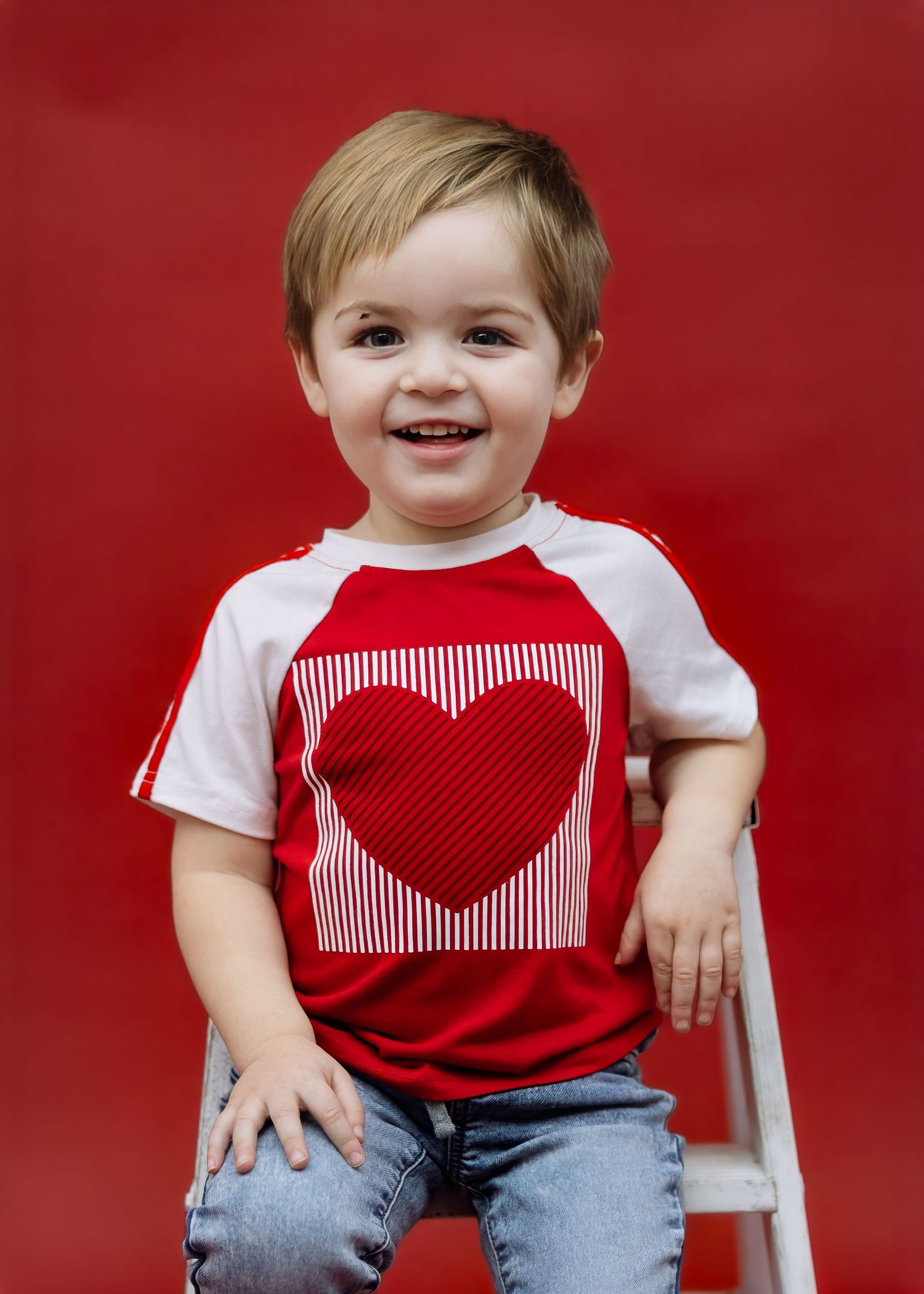 Smiling toddler boy in red and white heart raglan shirt sitting on white ladder stool.