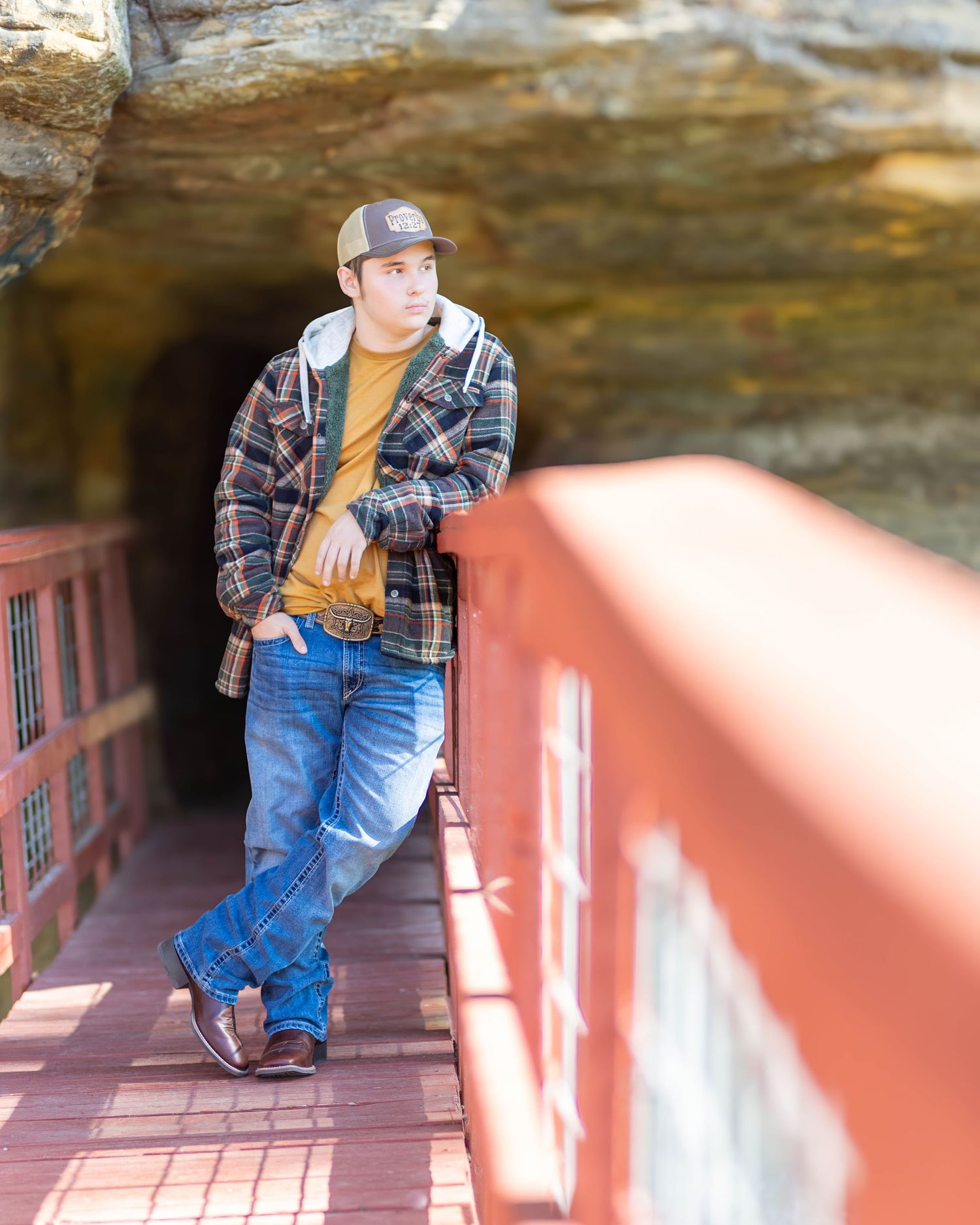 Teen boy in flannel jacket and trucker hat leans on red bridge railing for senior portrait.