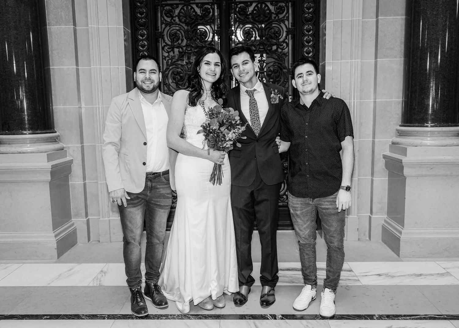 A bride and groom pose with two men in casual attire before ornate doors in a black and white photo.