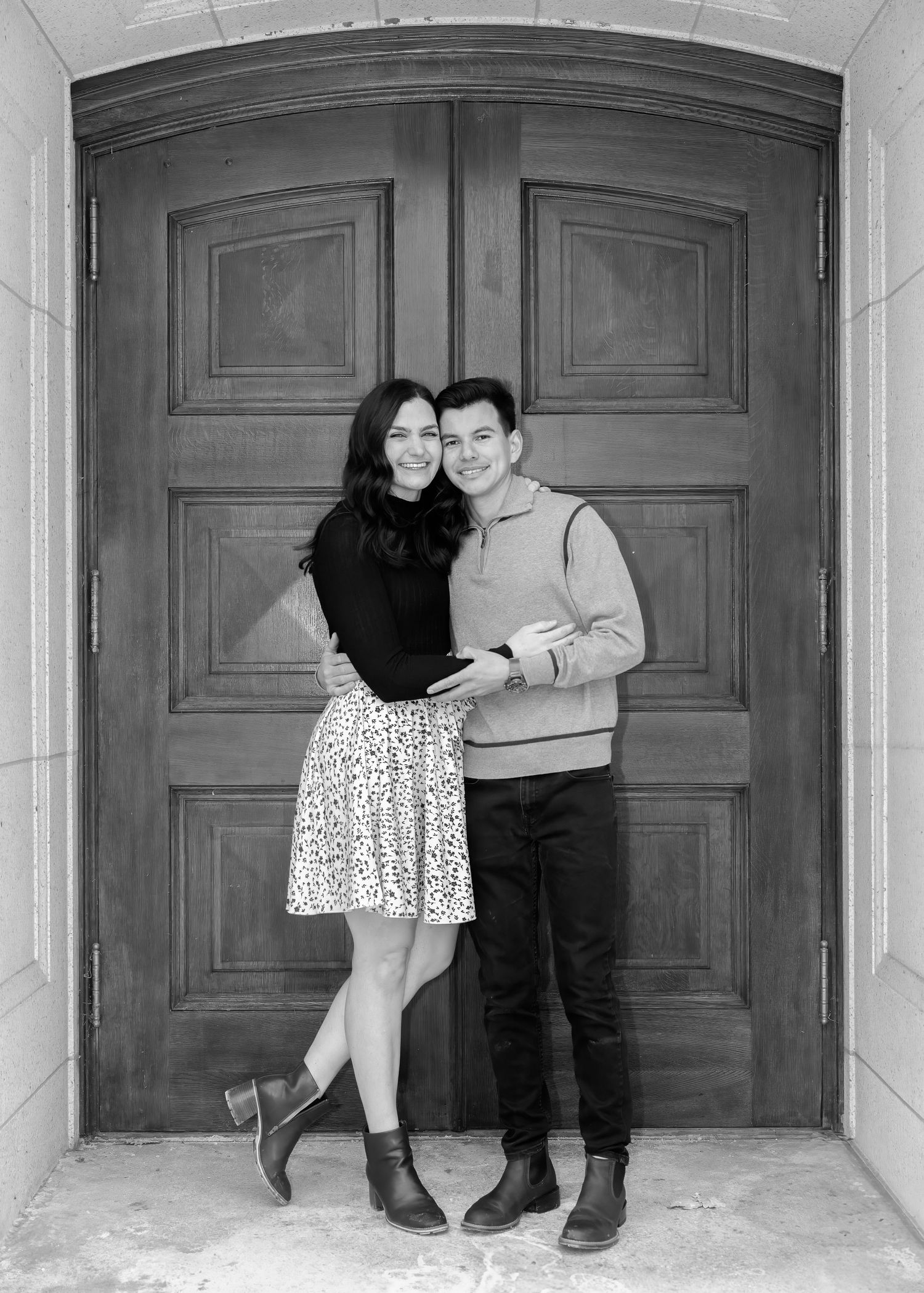 Black and white photo of couple embracing and smiling in front of large wooden double doors.