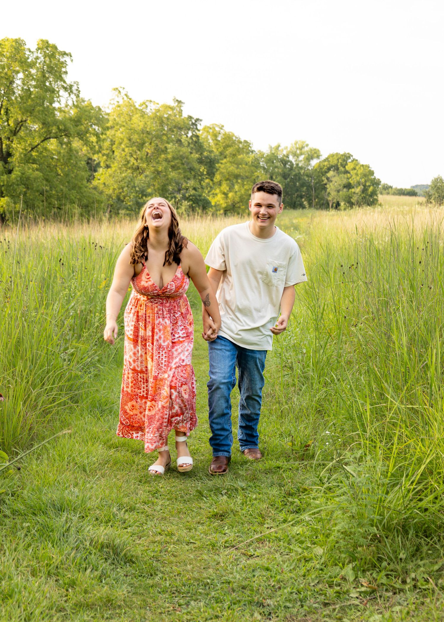A joyful couple laughs while walking hand-in-hand along a grassy path surrounded by tall green fields.