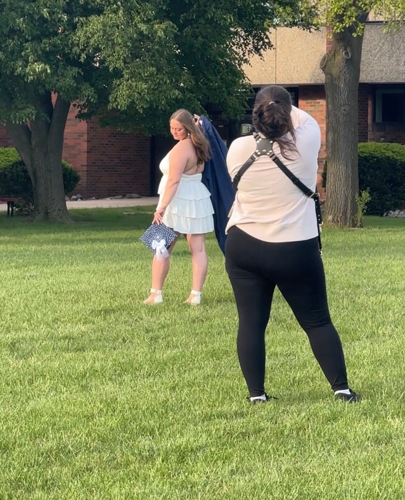 Two women outdoors on grass, one posing in a white skirt holding flowers while the other photographs her near a brick building.