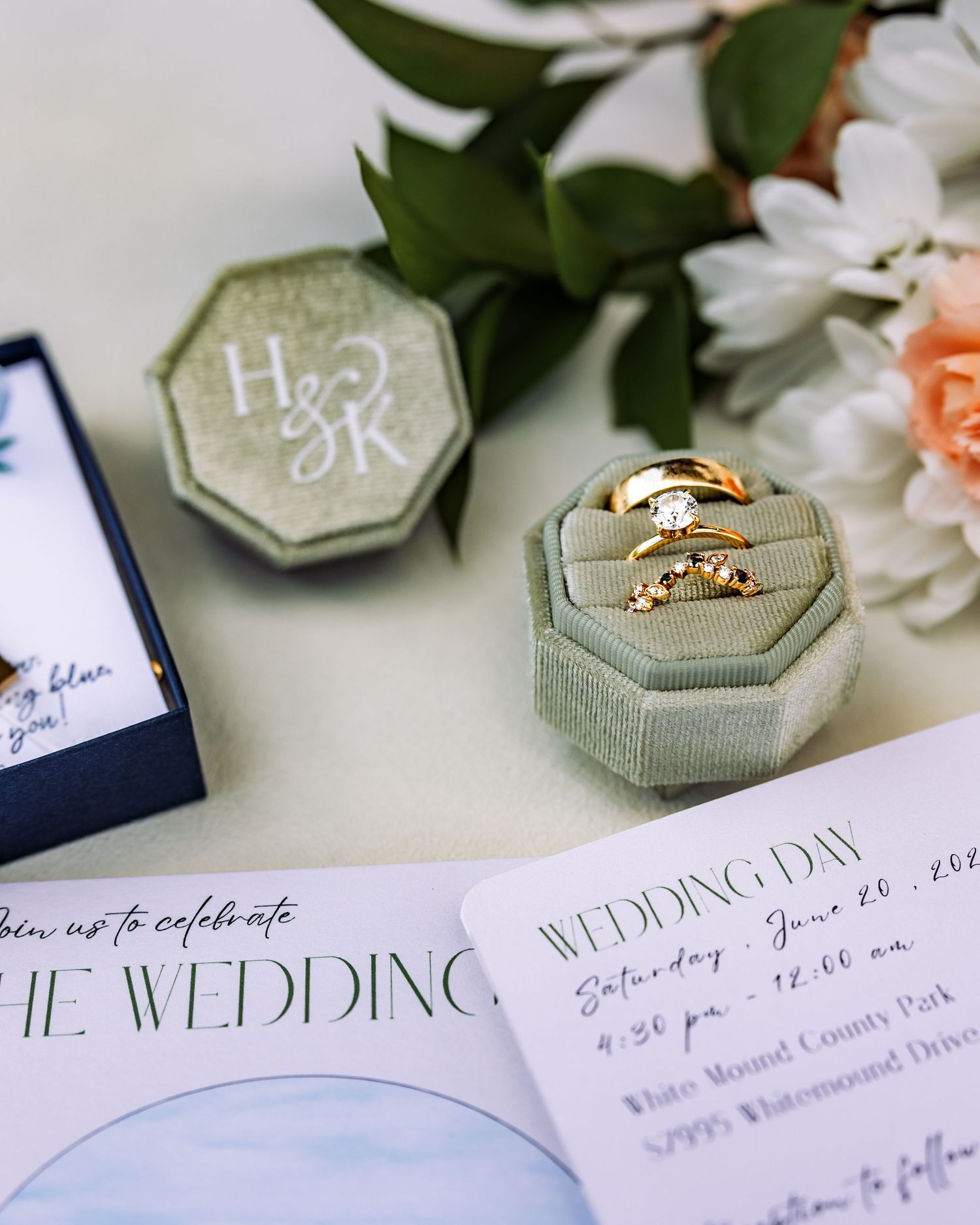 Gold wedding rings in a sage green velvet ring box with monogram H&K, beside floral wedding invitations and white flowers.