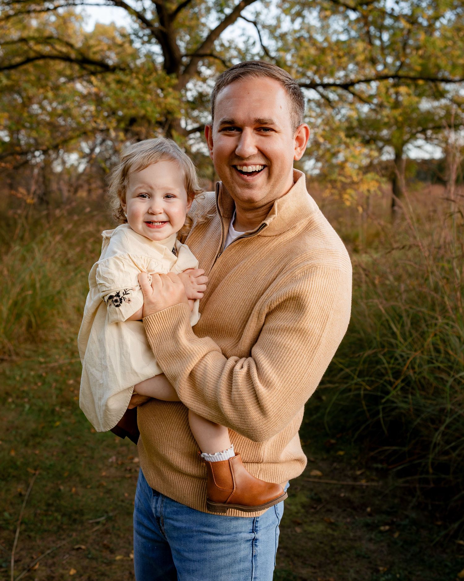 A smiling man in a tan sweater holds a laughing toddler girl outdoors among autumn trees.