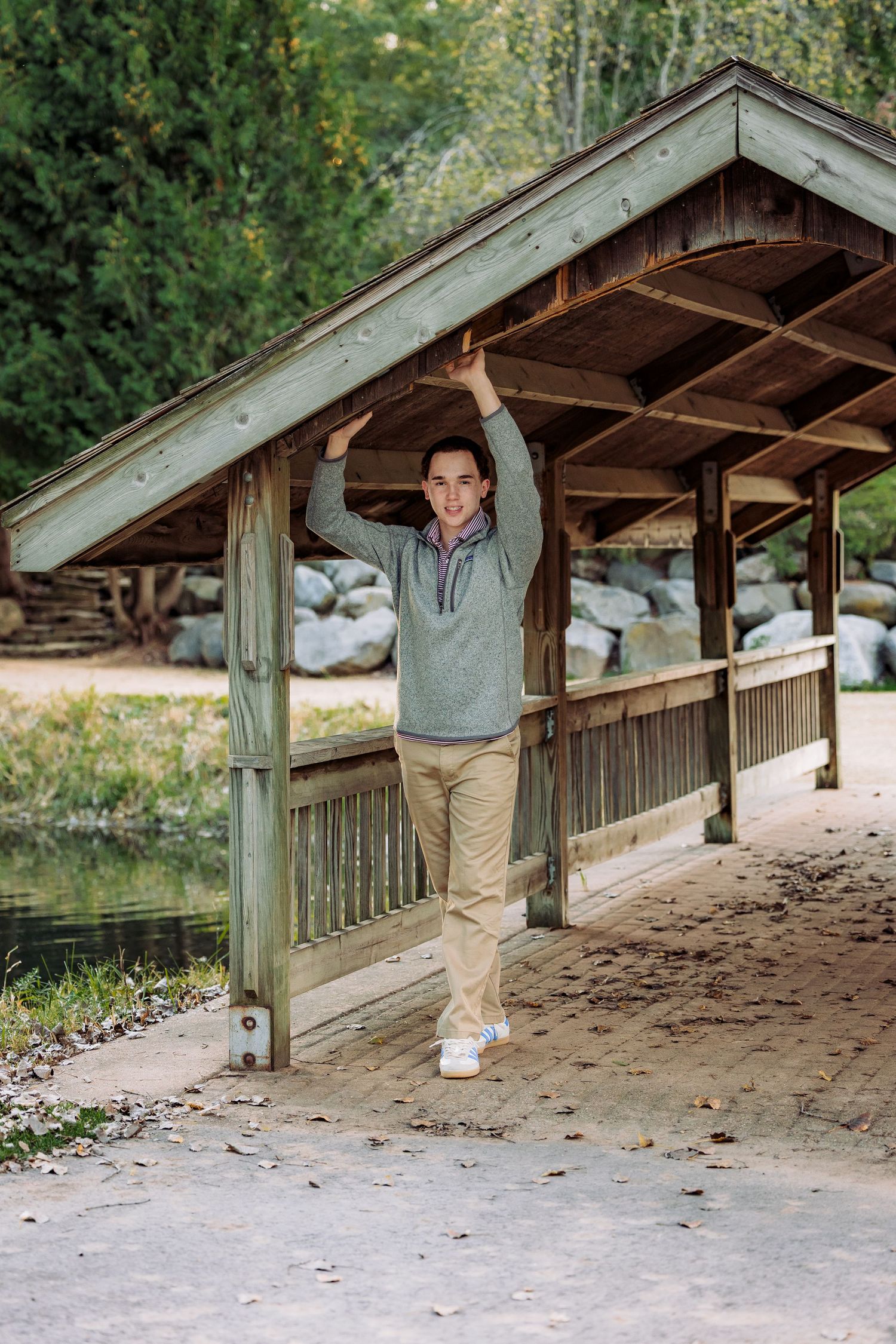 Teen boy in gray sweater and khaki pants poses under a wooden covered bridge in a scenic outdoor park setting.